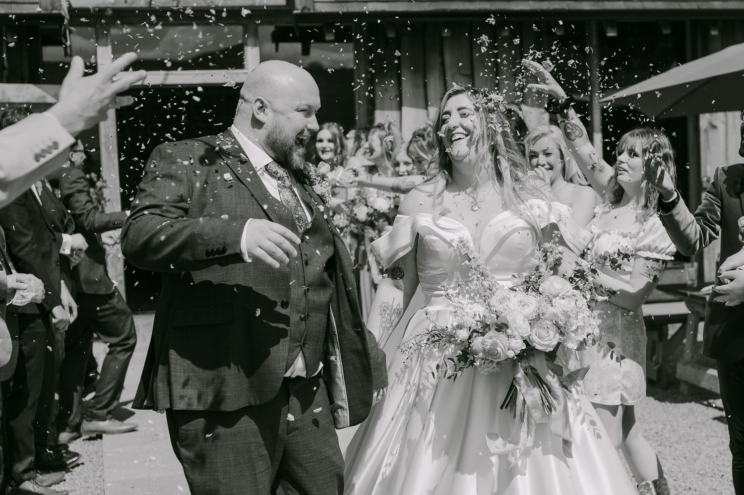 Black and white photo of bride and groom celebrating with confetti shower outside wedding venue.
