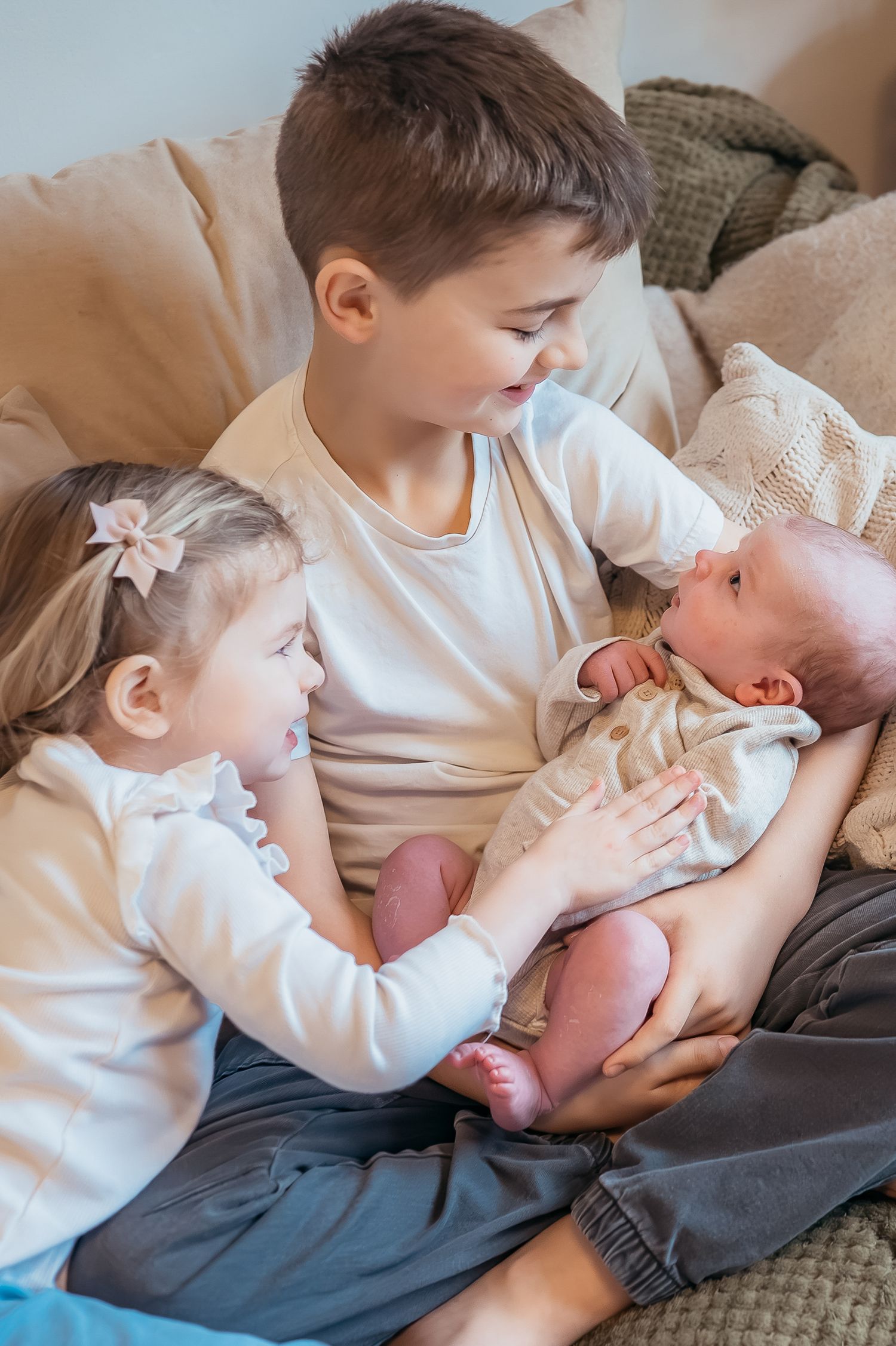 Two children lovingly hold their newborn sibling on a cozy couch, capturing a precious family moment.