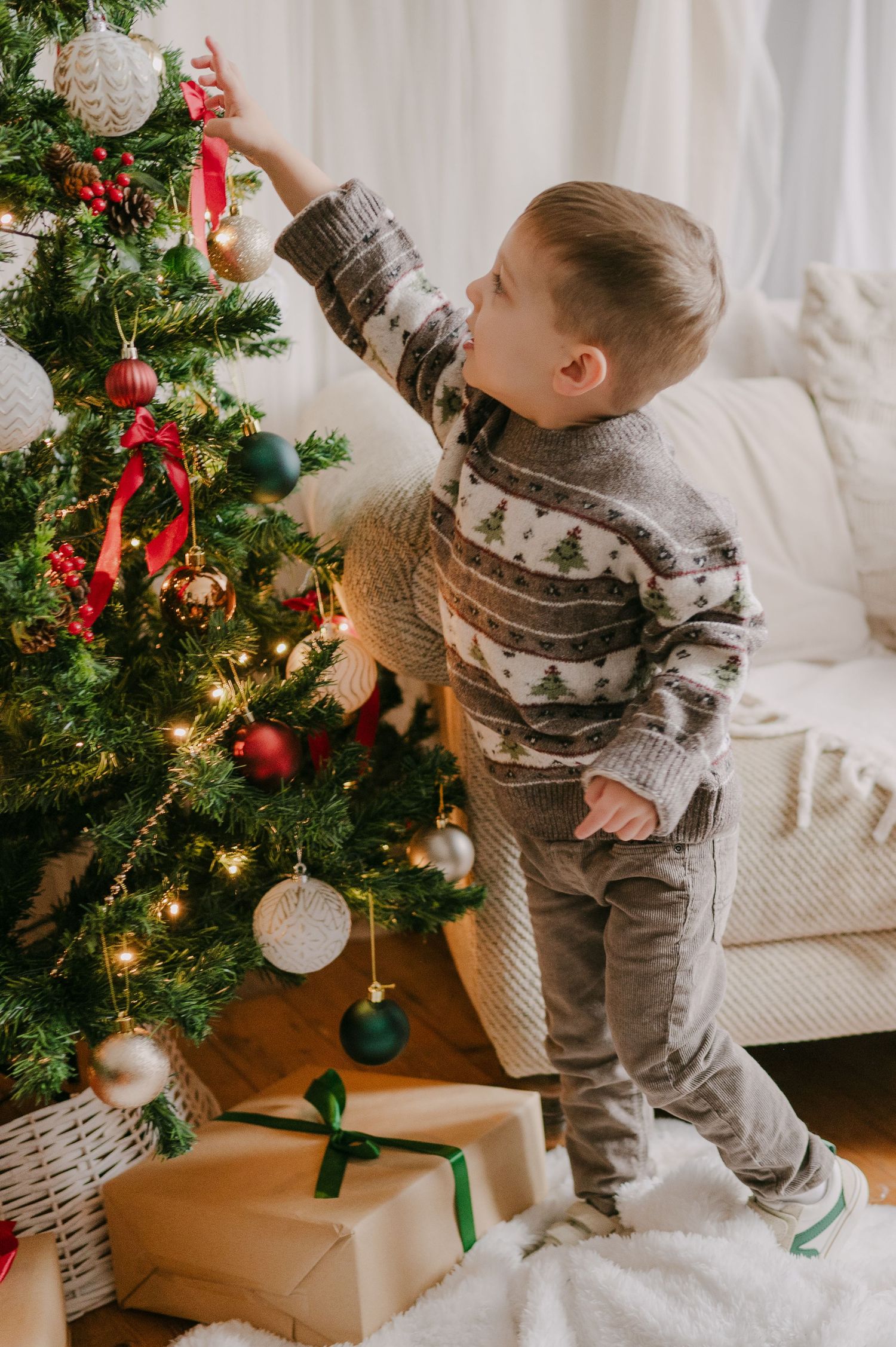 Young boy in a Christmas tree patterned sweater reaching to decorate a lit holiday tree with ornaments.