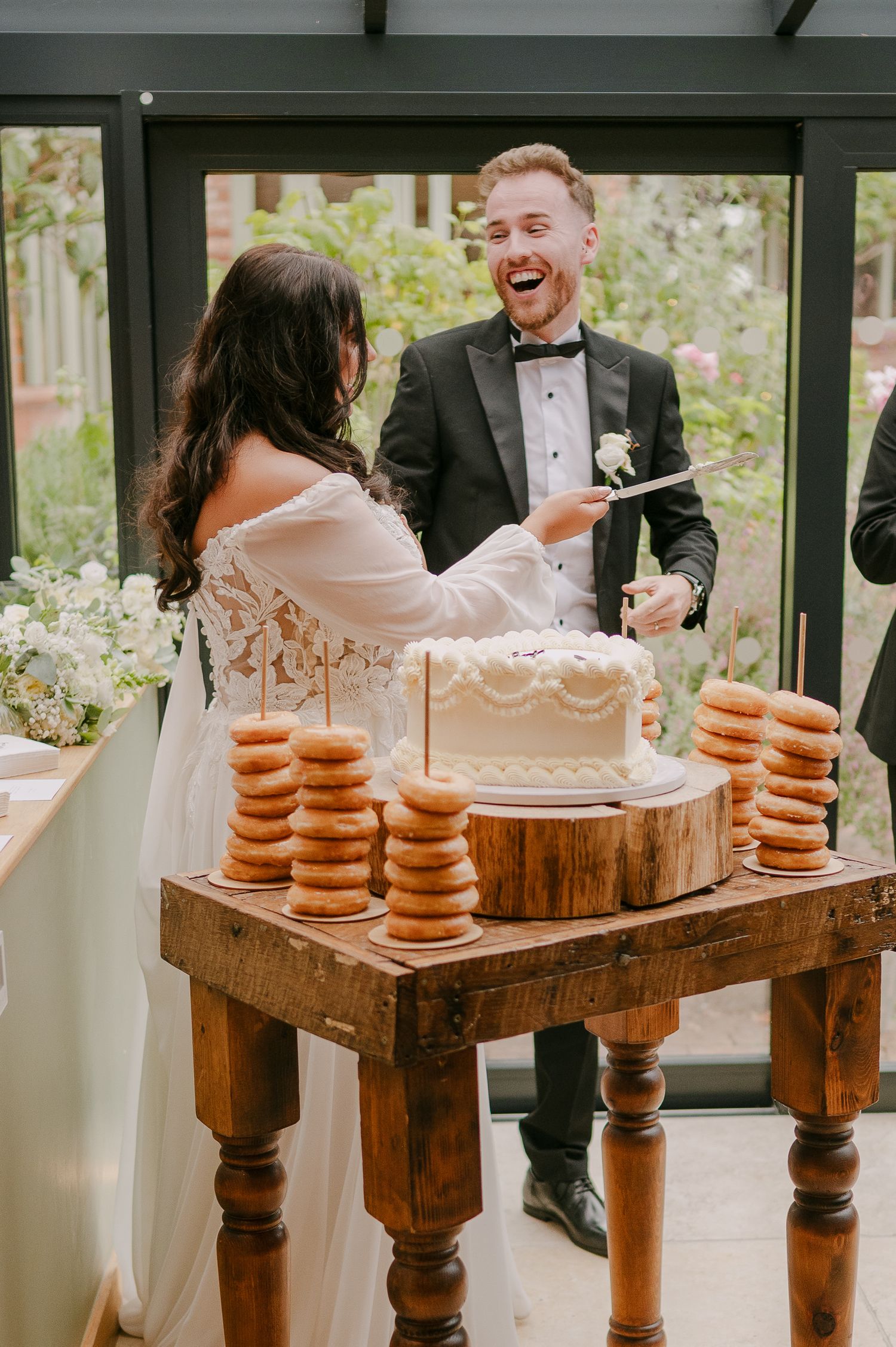 Laughing groom watches bride cut wedding cake surrounded by stacked donuts on rustic wooden table.
