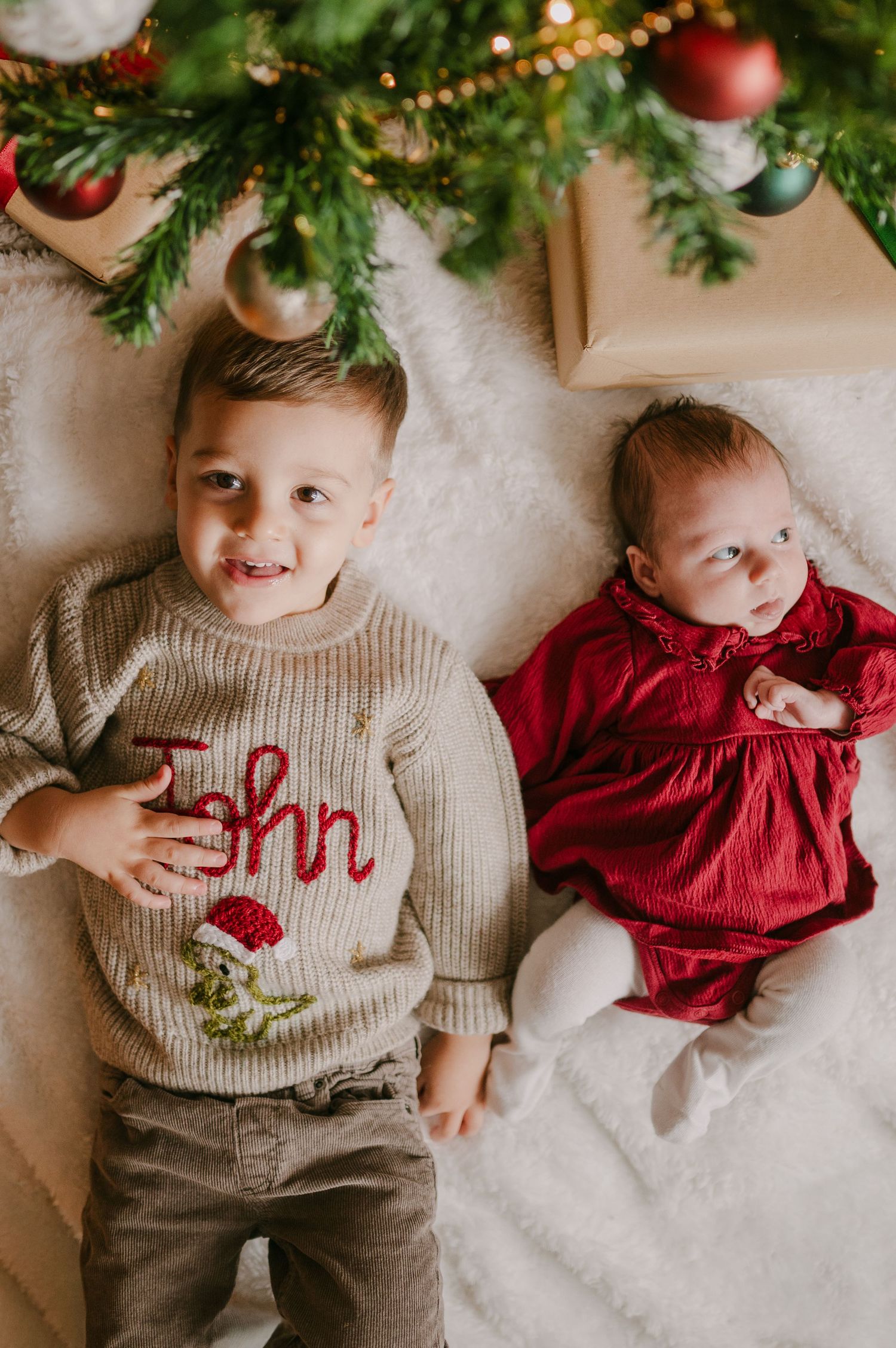 Toddler boy in personalized John Christmas sweater lying beside baby girl in red dress under holiday tree.