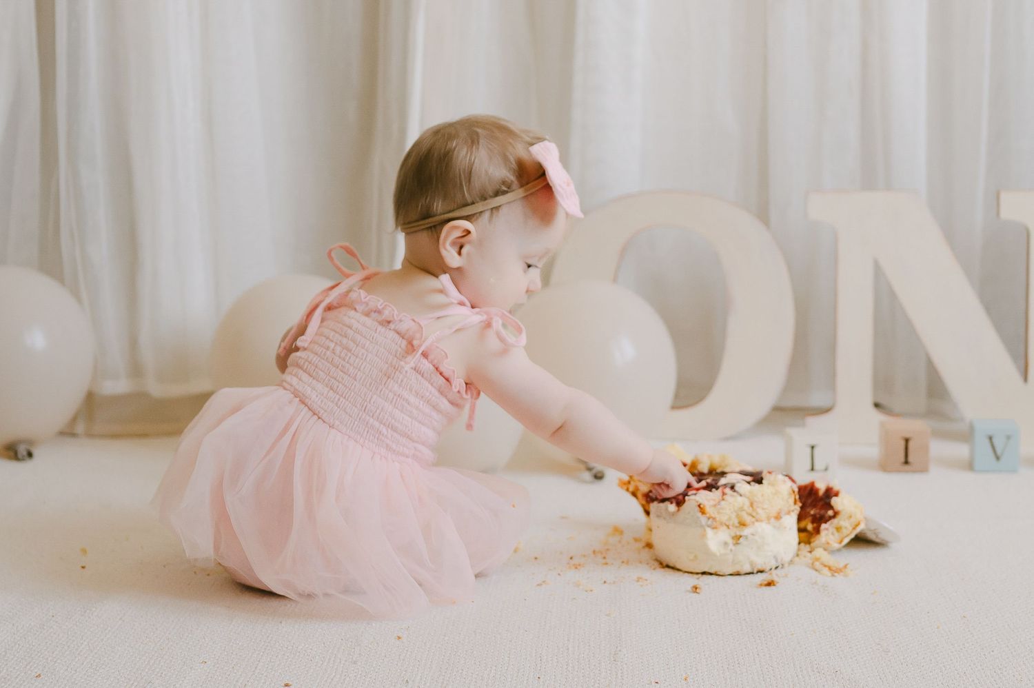 Baby girl in pink dress reaching into her birthday cake during a first birthday cake smash session.