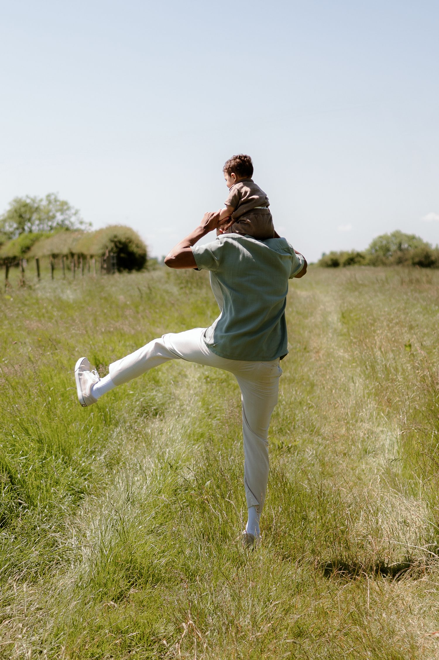 Father carrying child on shoulders while playfully kicking leg in sunny green field.