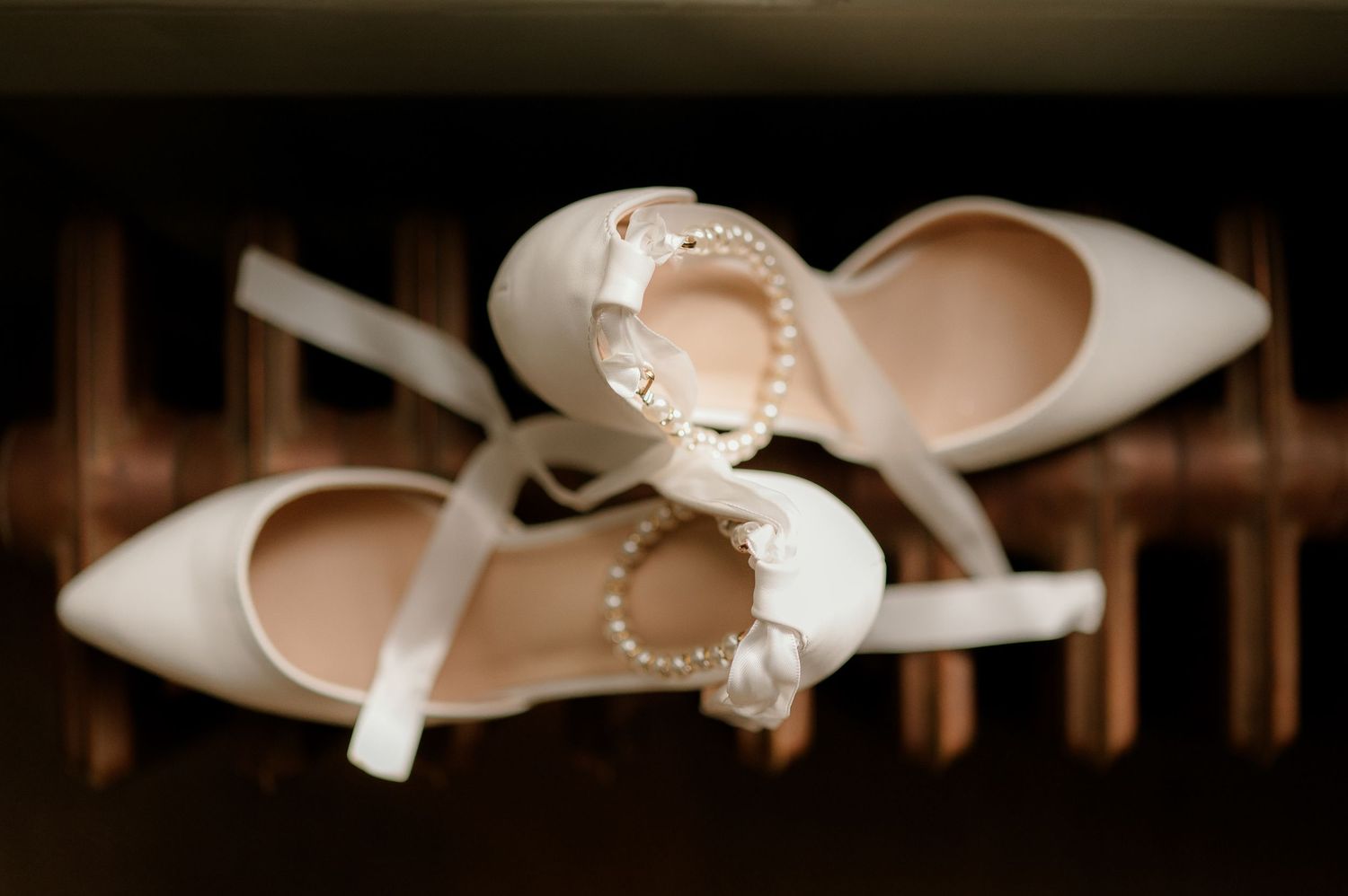 White bridal heels with pearl ankle straps and bow detail displayed on dark reflective surface.
