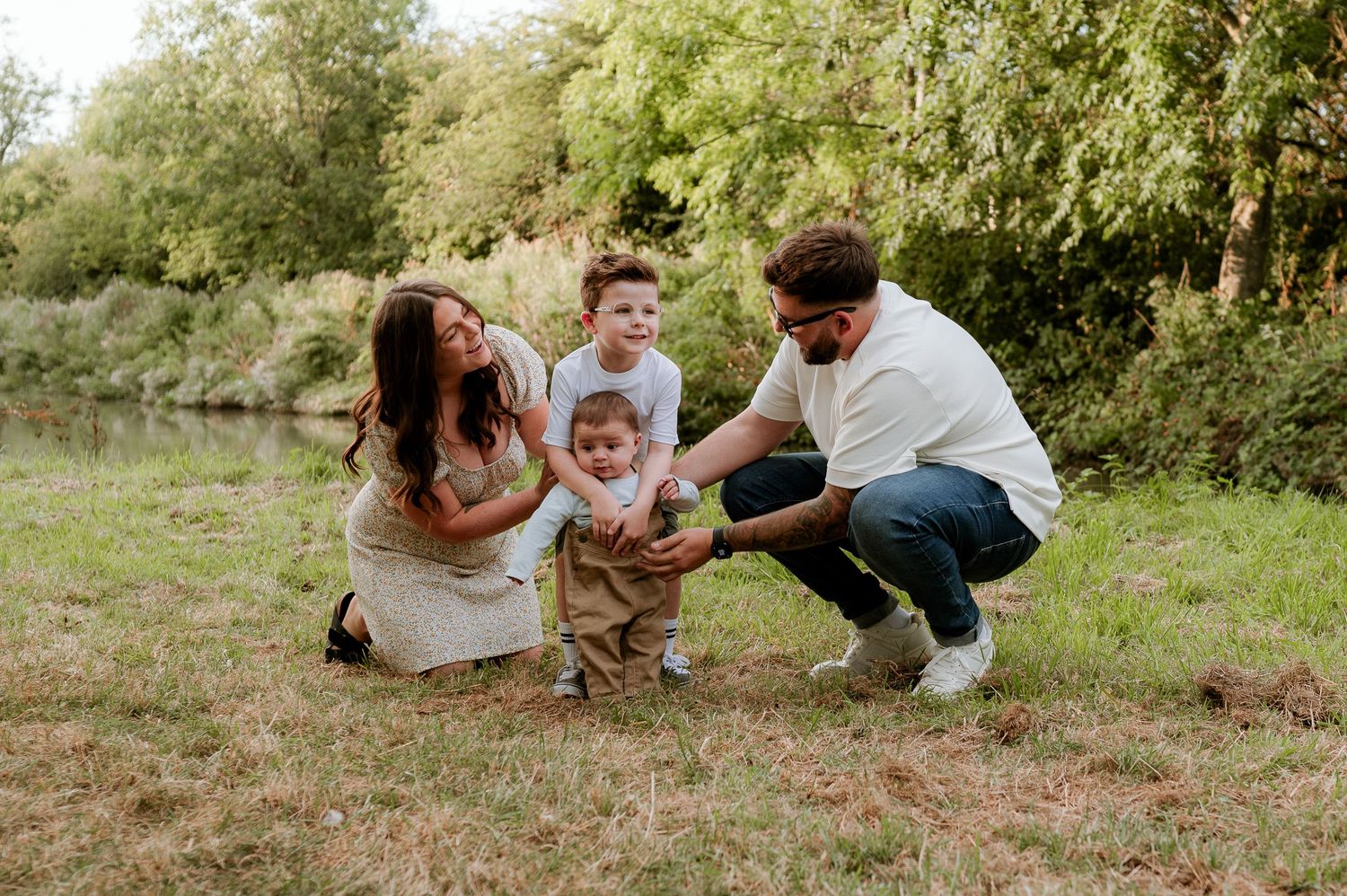 Family of four enjoying outdoor photos near a pond, parents helping toddler walk while older child stands nearby.