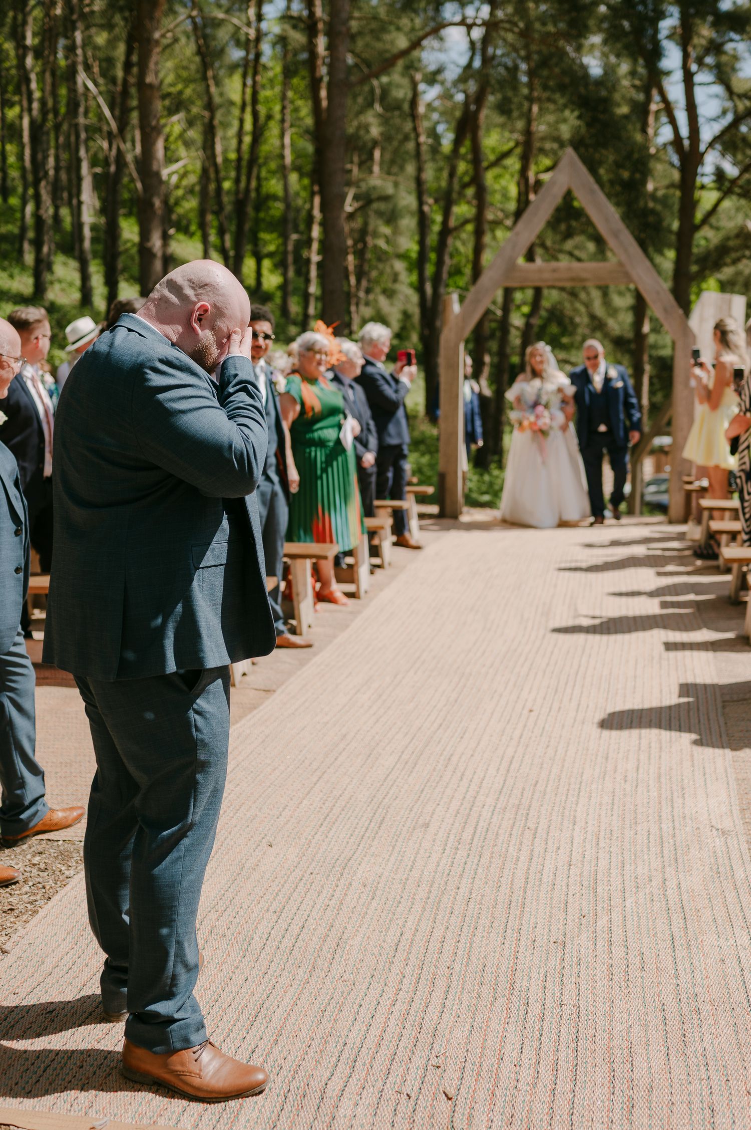 Emotional bald wedding guest wipes tears as bride walks down outdoor woodland aisle ceremony.