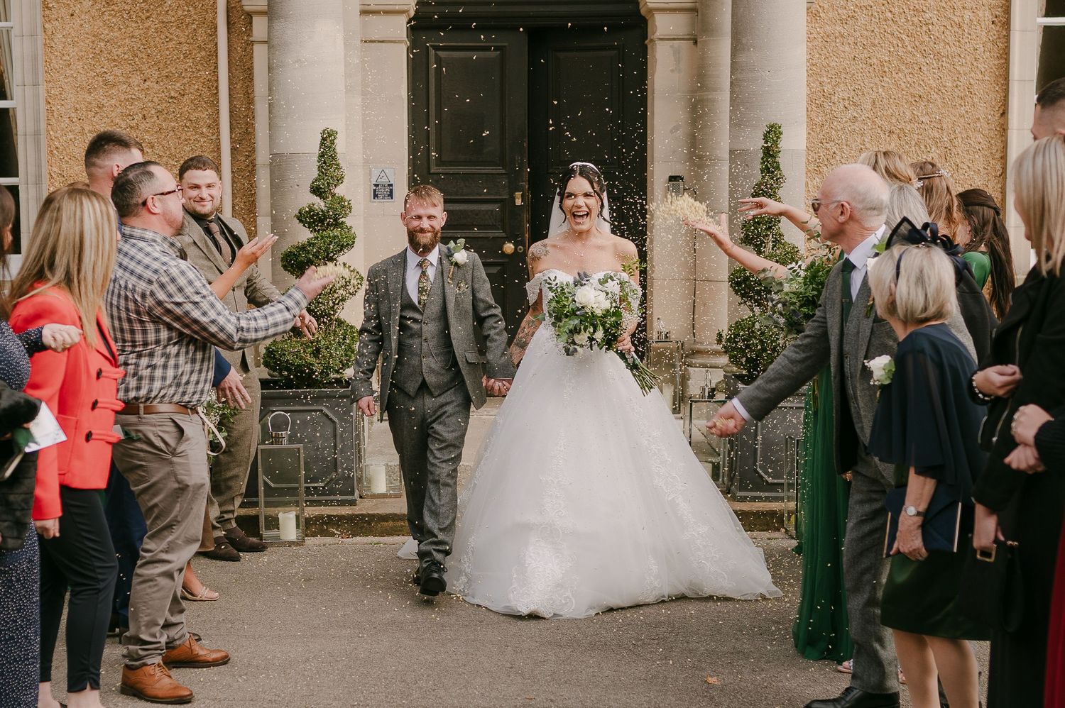 Newlywed couple walking through confetti tunnel with guests celebrating outside an elegant country house venue.