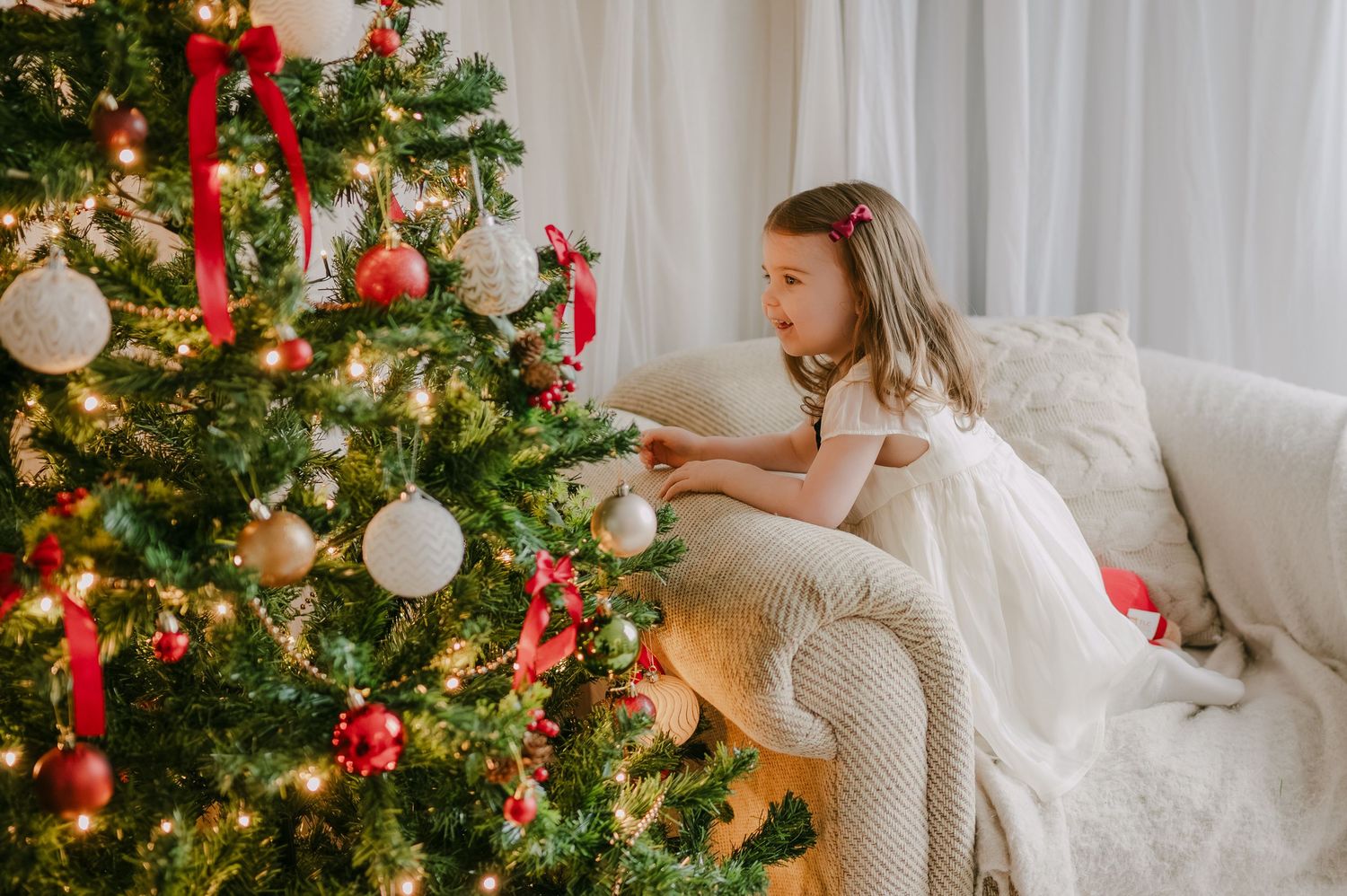 Little girl in white holiday dress sitting on sofa reaching to touch ornaments on a Christmas tree.