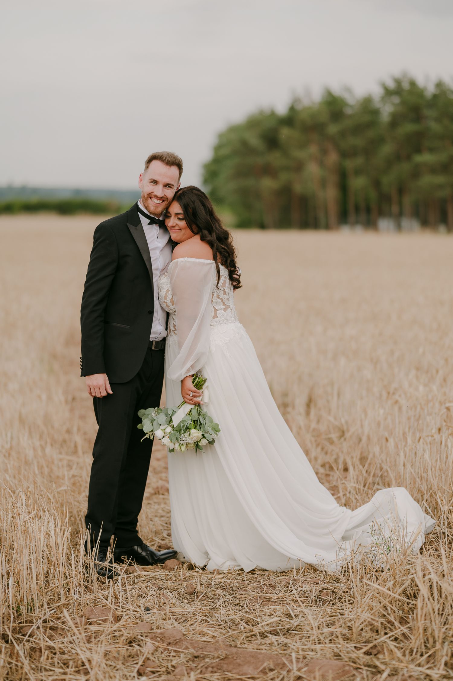 Bride and groom embracing romantically in a golden wheat field with pine trees in background.