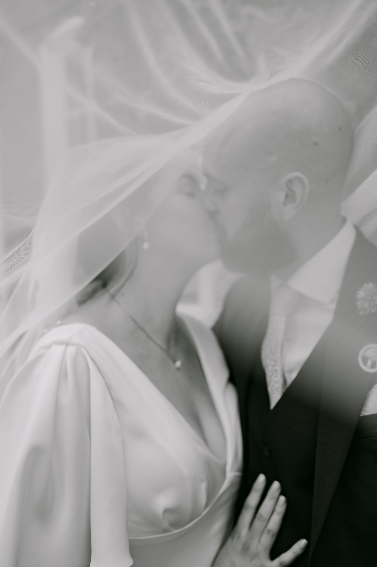 Black and white photo of bride and groom kissing under a flowing wedding veil.