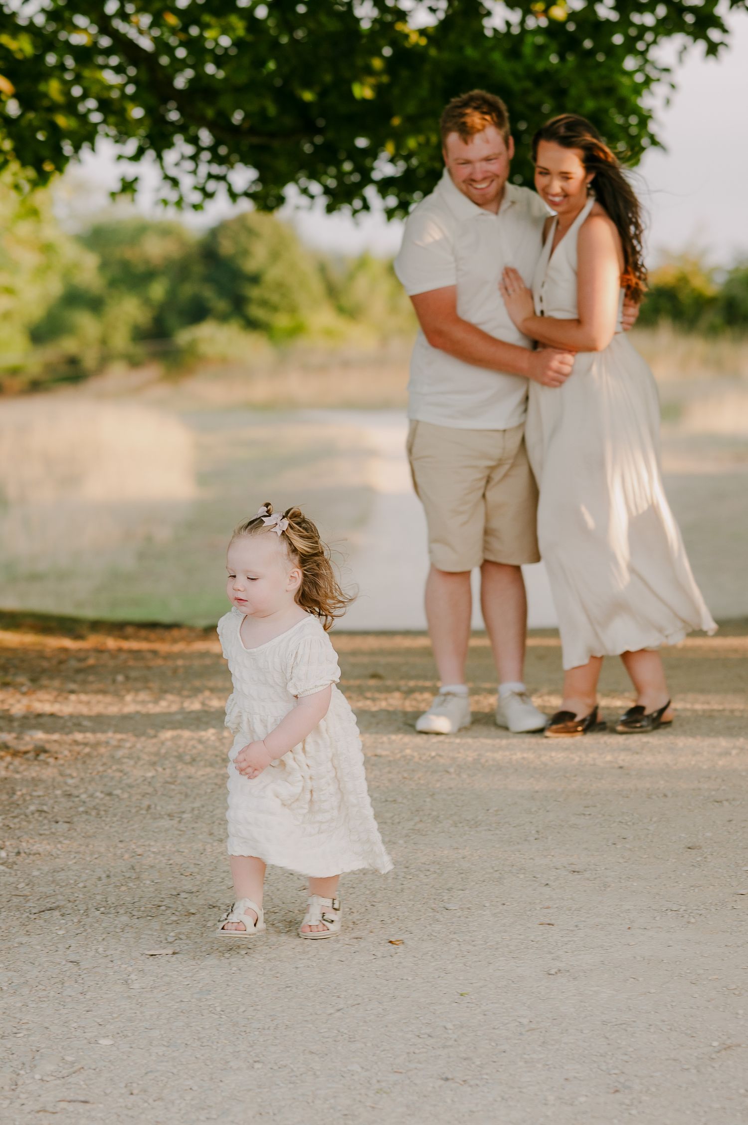 Toddler girl in white dress walking independently as her parents laugh together in golden sunlight.