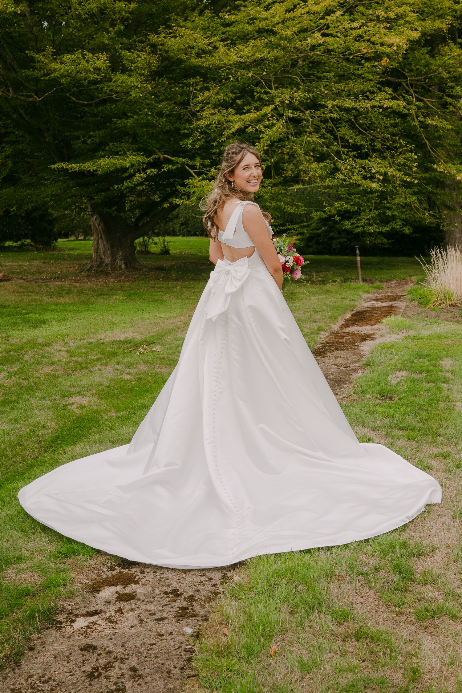Bride in stunning white ballgown with large bow back and long train poses in lush green garden setting.