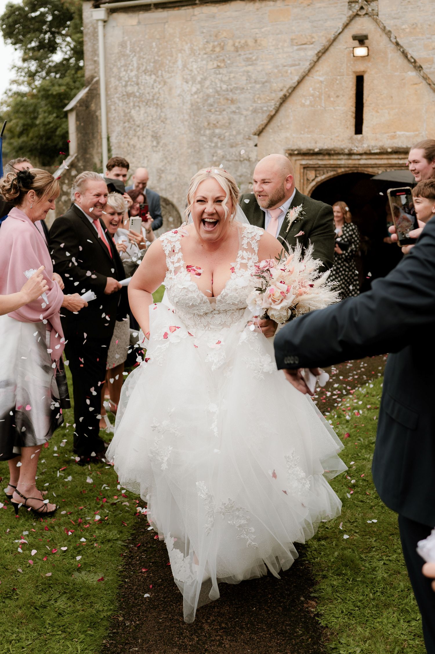 Joyful bride in layered tulle gown laughs as guests throw pink confetti outside stone church.