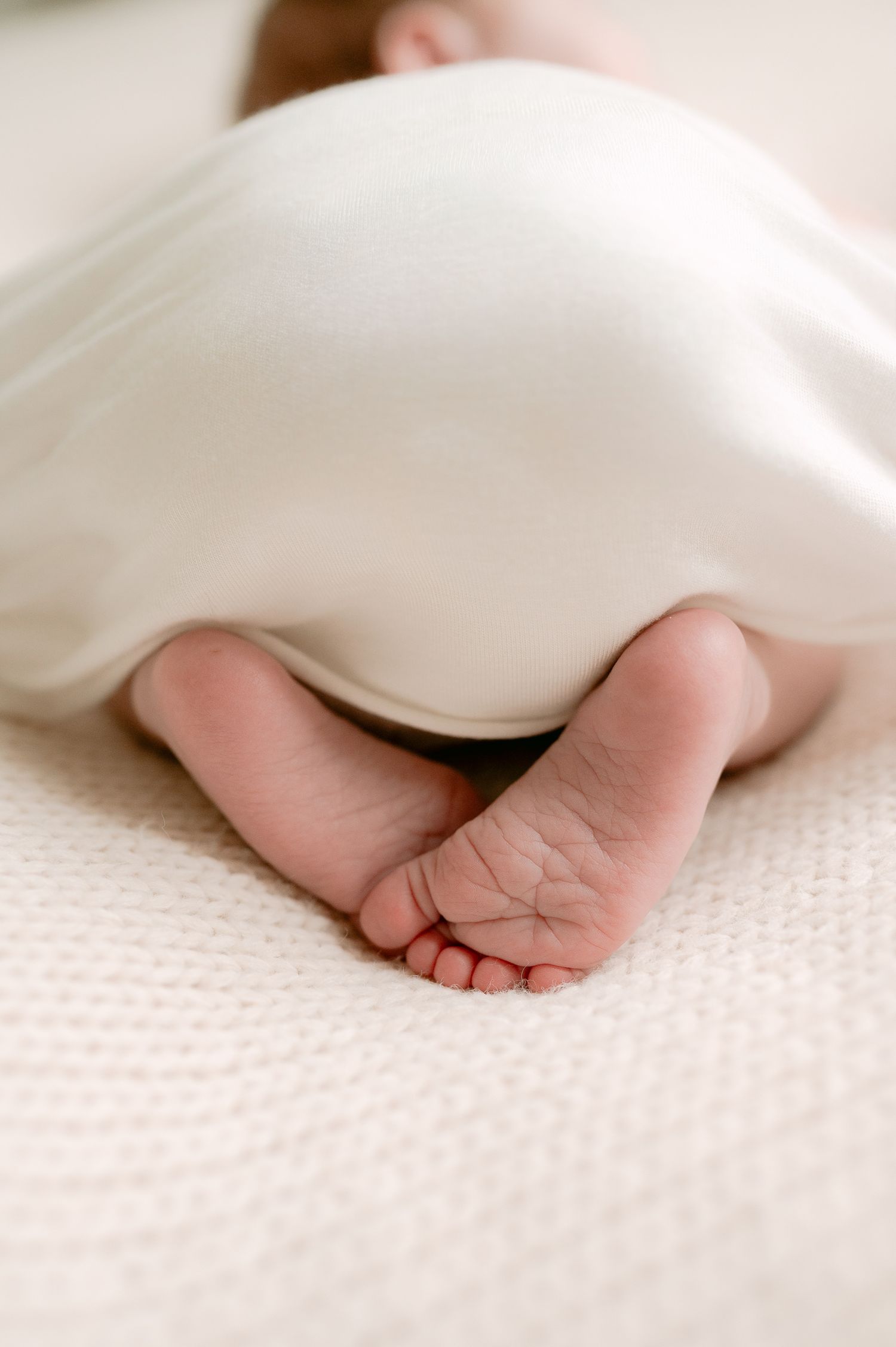 Newborn baby feet peeking out from under a white silk blanket on a cream textured surface.