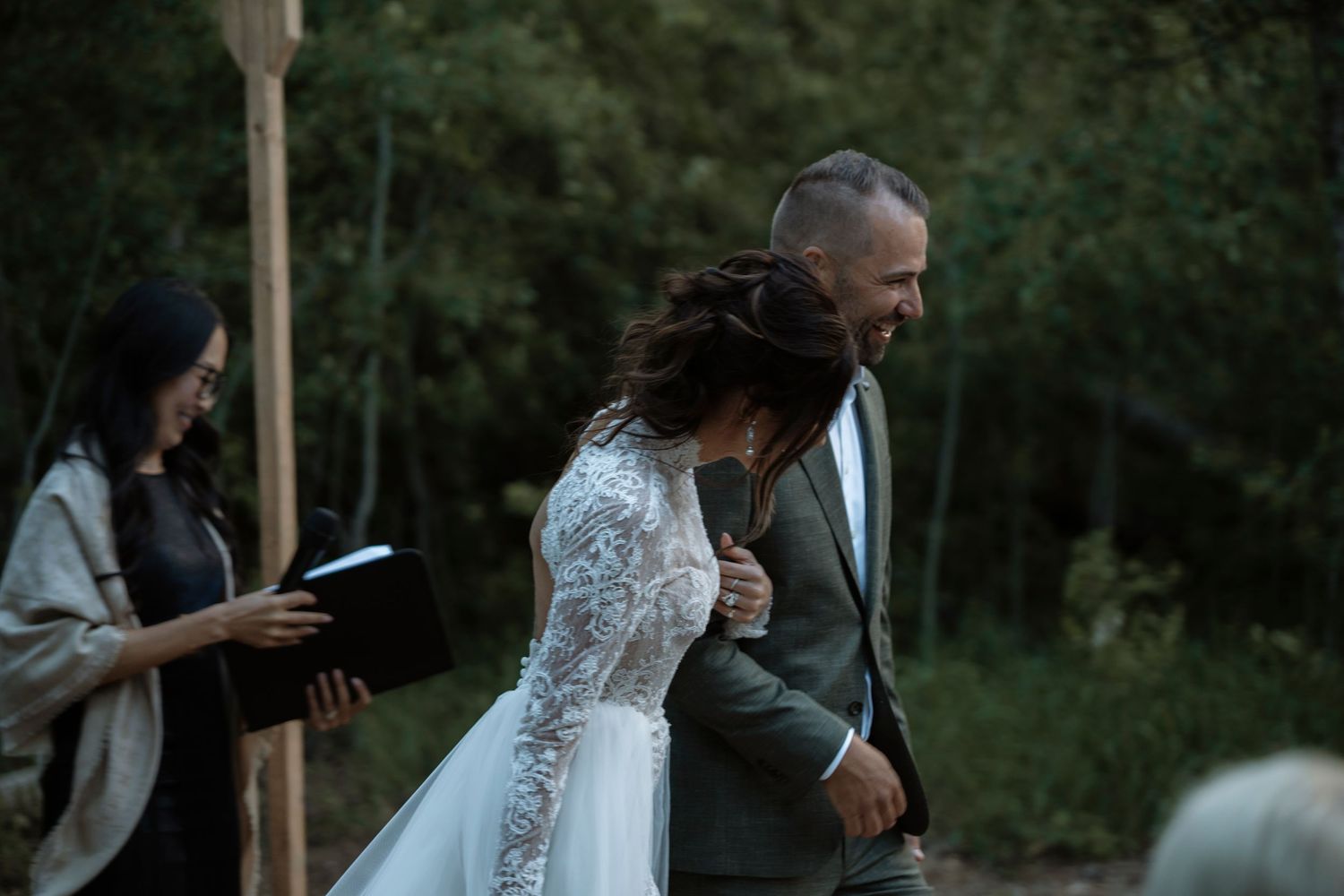 Bride and groom share a joyful moment during outdoor wedding ceremony with officiant holding book in forest setting.