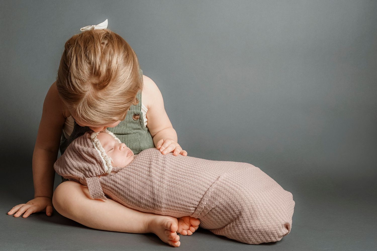 Young toddler girl in green outfit tenderly leaning over a pink-swaddled sleeping newborn baby.Family photography in Regina Saskatchewan.