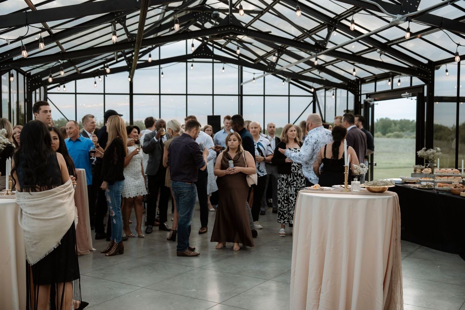 Guests mingle at an elegant indoor greenhouse venue reception with high glass ceilings and cocktail tables.
