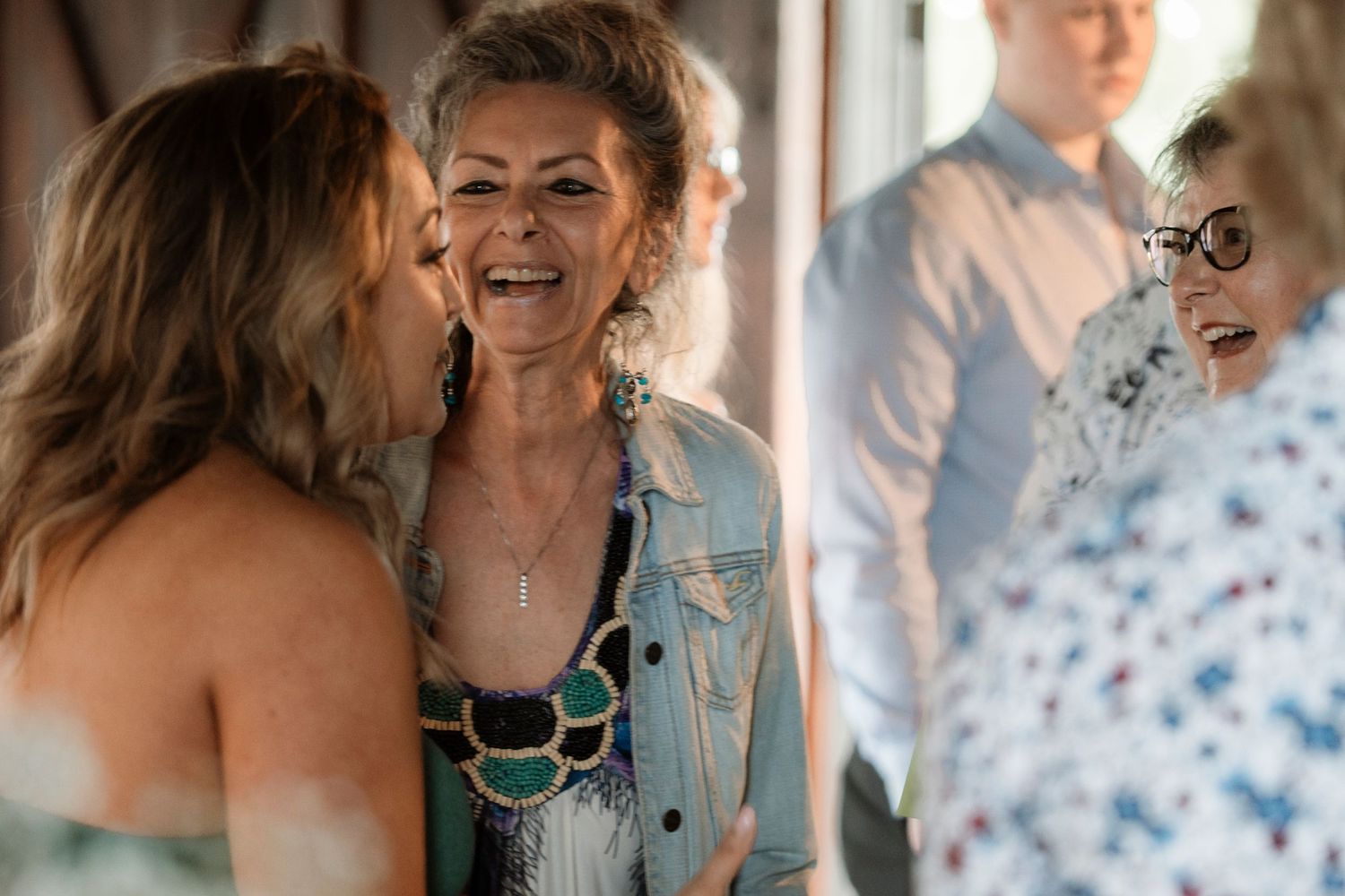 Women laughing and sharing a joyful moment at a social gathering, with guests visible in the background.