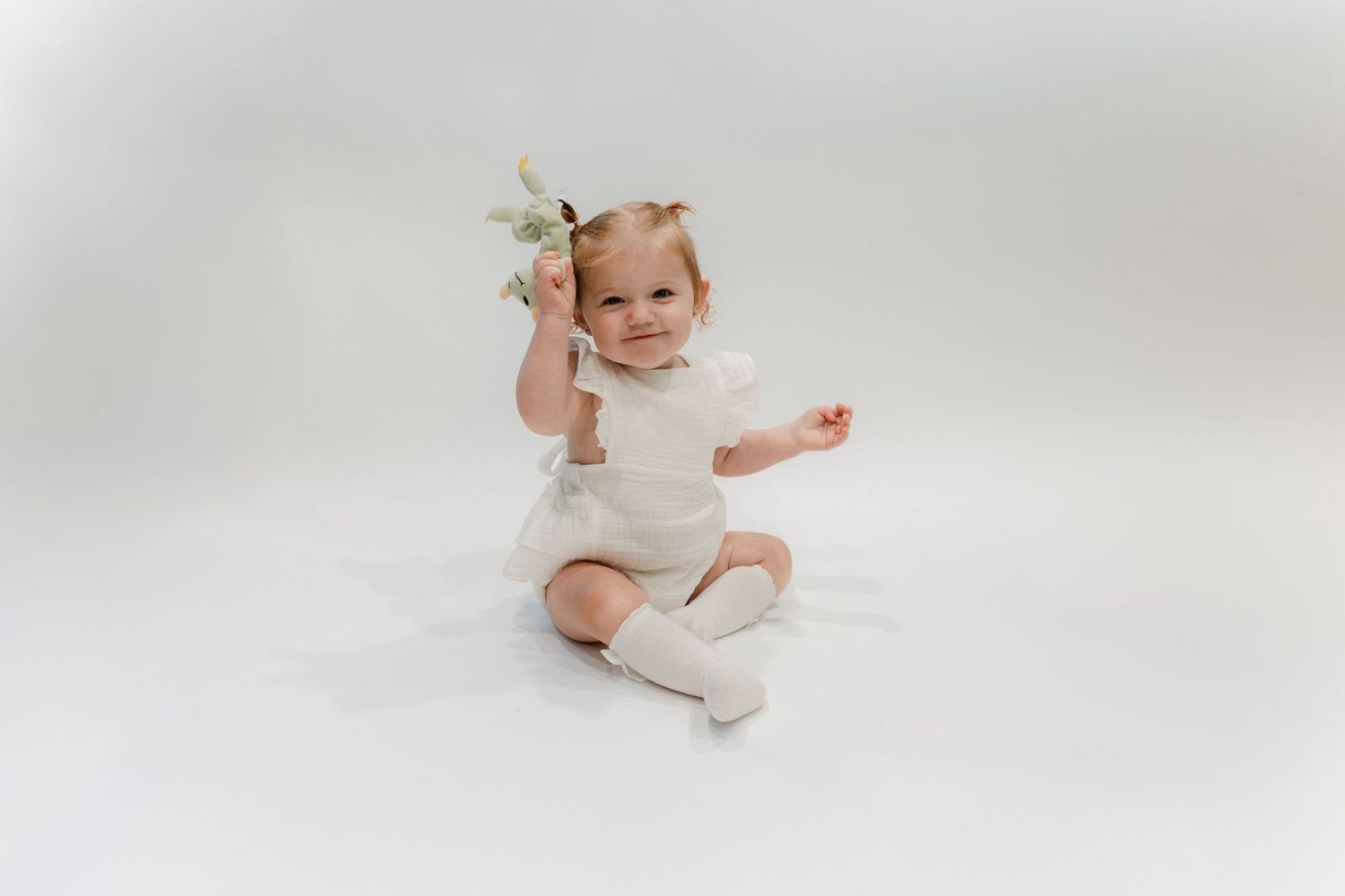 Adorable baby girl in white outfit sitting on floor holding a small flower bouquet.Family photography in Regina Saskatchewan.