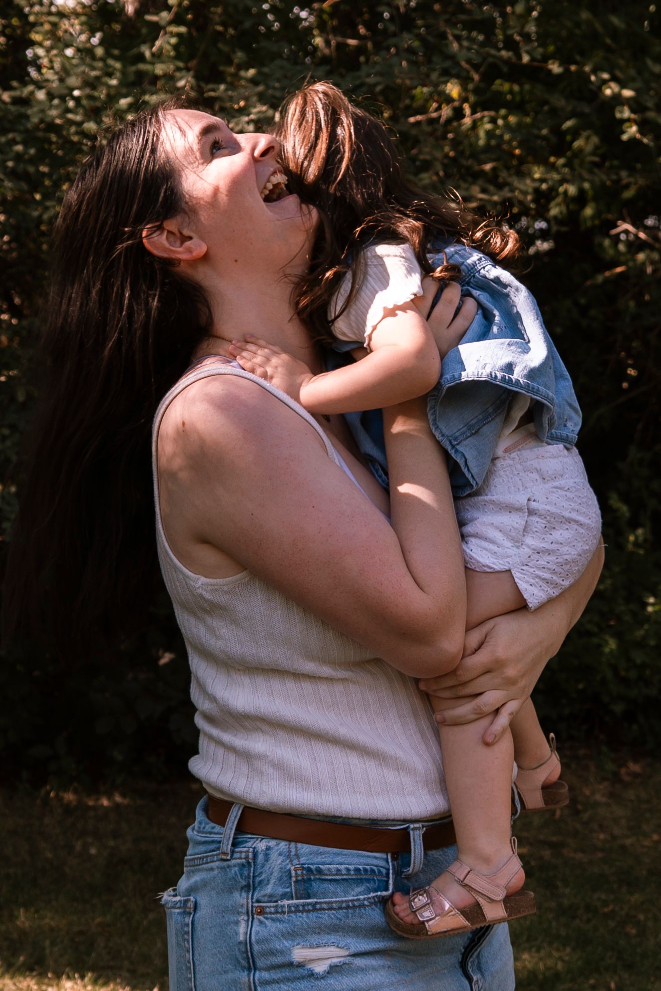 A laughing mother holds her toddler up close in a sunny outdoor garden setting.Family photography in Regina Saskatchewan.