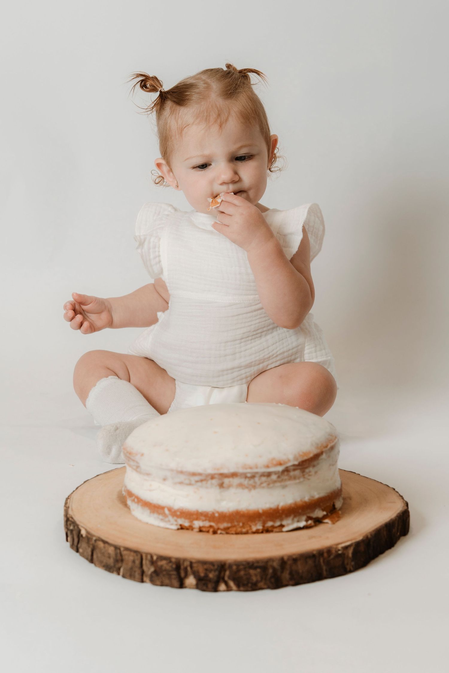 A baby girl in a white outfit sits on a wood slice tasting her first birthday smash cake.Family photography in Regina Saskatchewan.