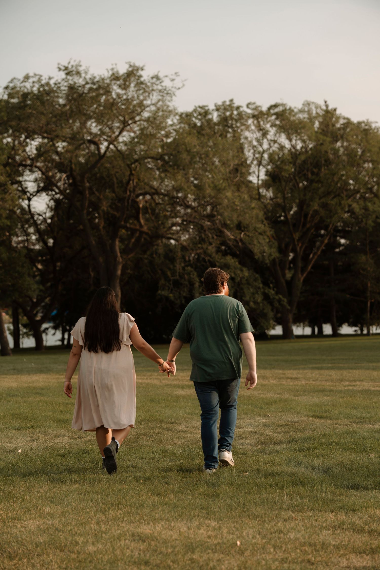 Couple holding hands walking through a lush green park at golden hour, woman in white dress, man in green shirt.
