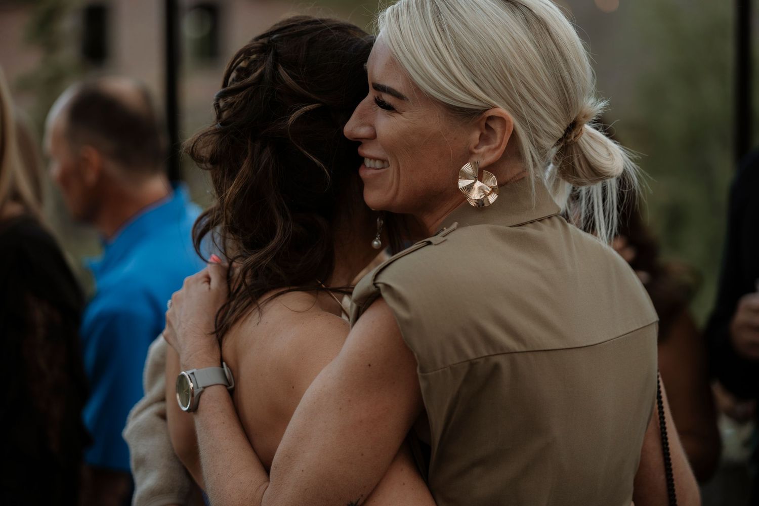 Two women share a warm embrace at an outdoor wedding reception, one wearing elegant pearl earrings.