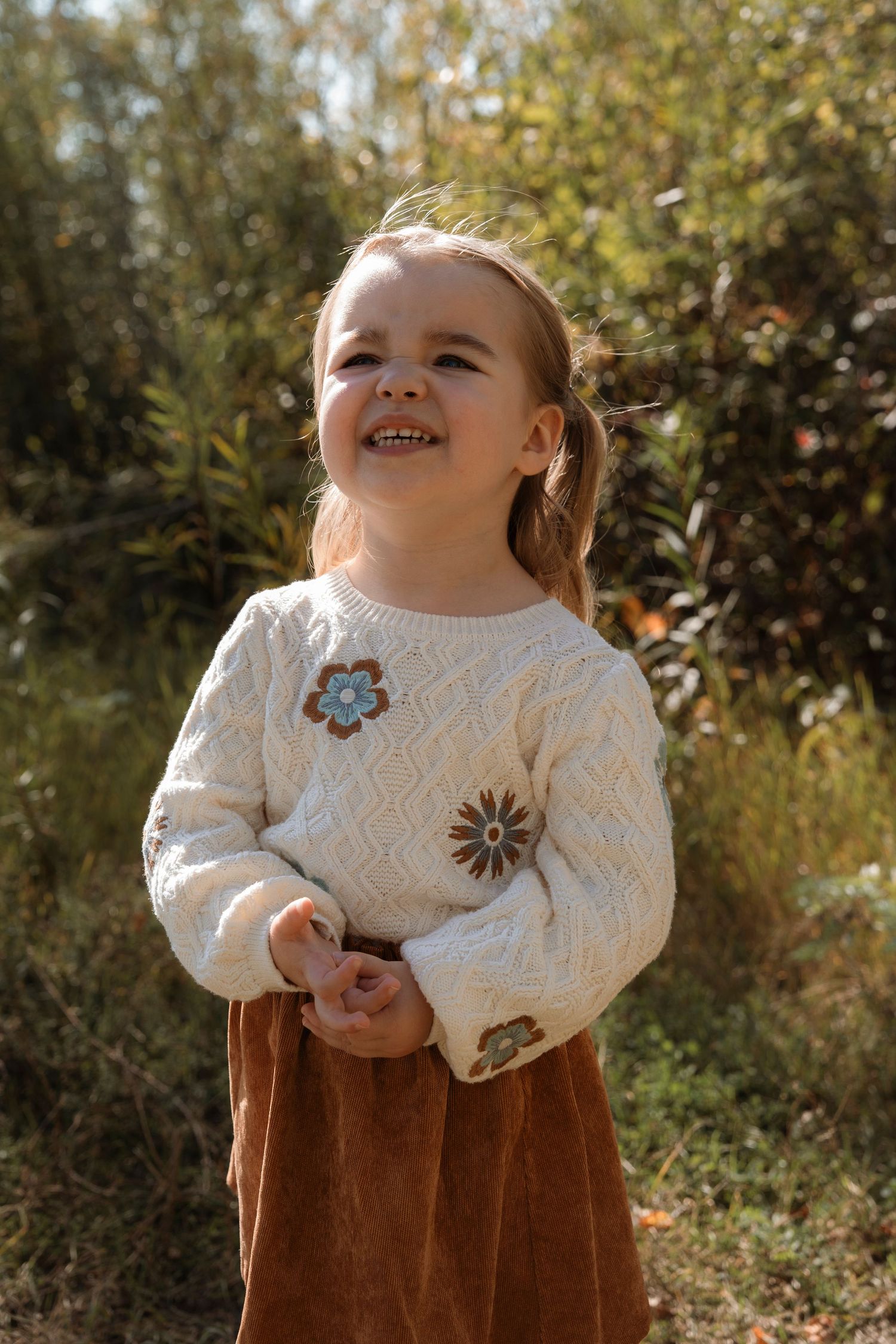 A happy young girl in a cream floral sweater and rust skirt smiles while standing in nature.Family photography in Regina Saskatchewan.