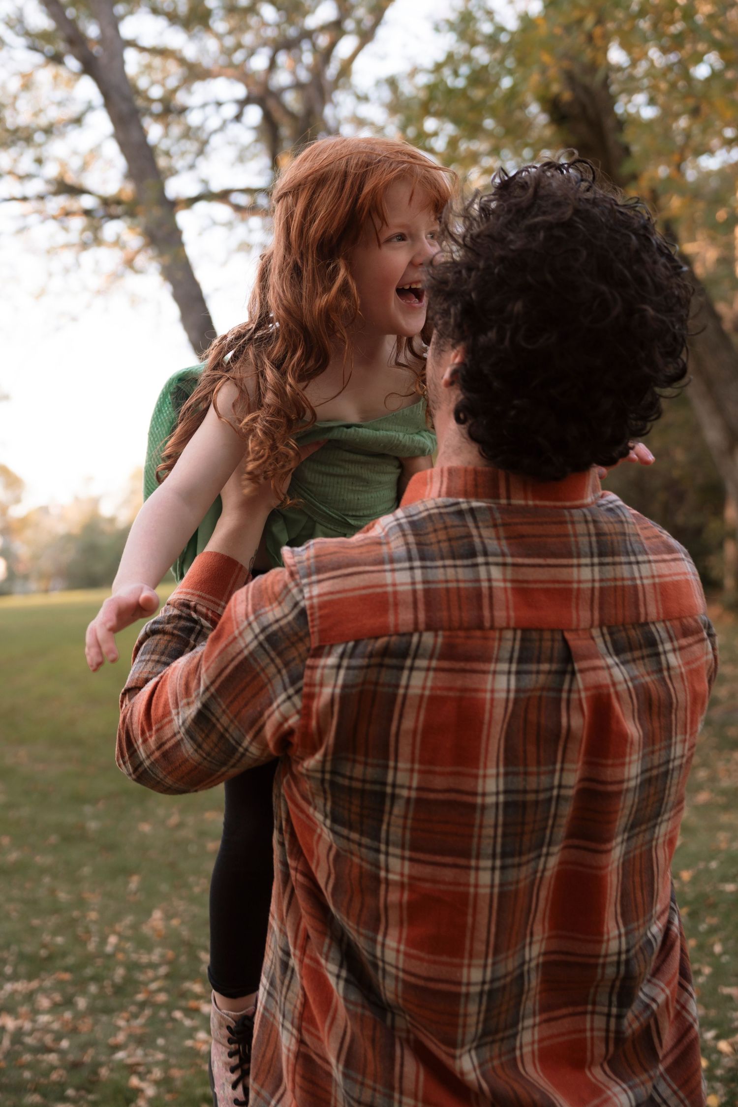 A laughing red-haired girl is lifted by her curly-haired father in an autumn park setting. Family photography in Regina Saskatchewan.