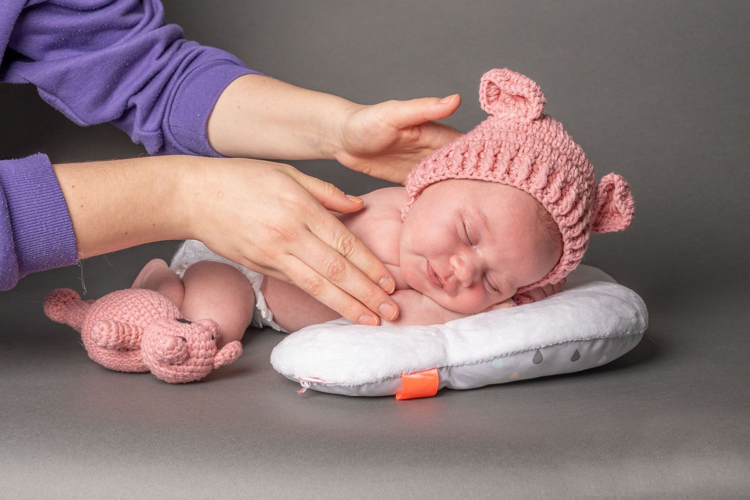 Newborn baby sleeping on white pillow wearing pink knit hat with adult hands gently cradling head beside crochet toy.