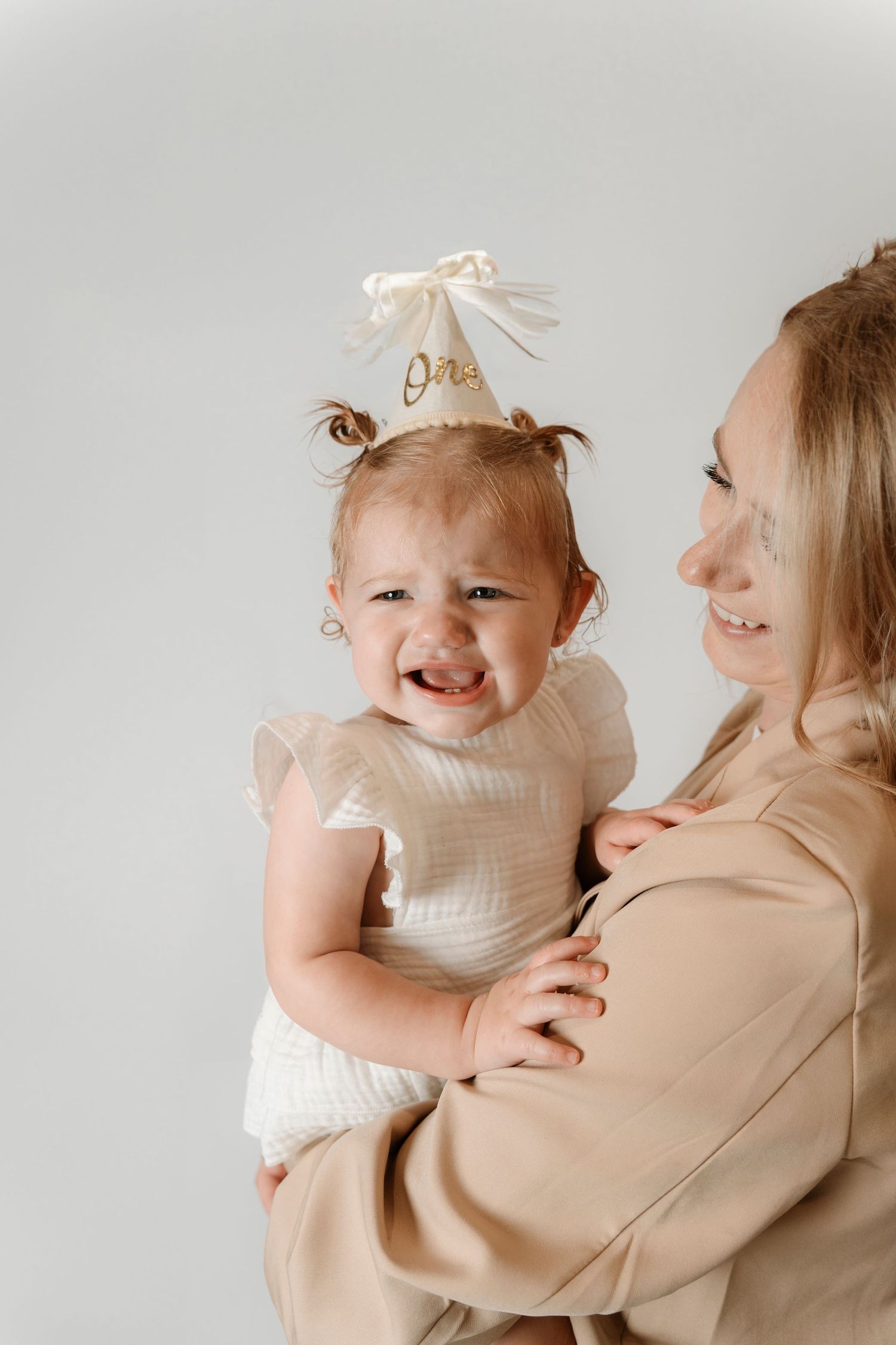 A baby girl wearing a One birthday crown cries while being held by a smiling blonde woman.Family photography in Regina Saskatchewan.