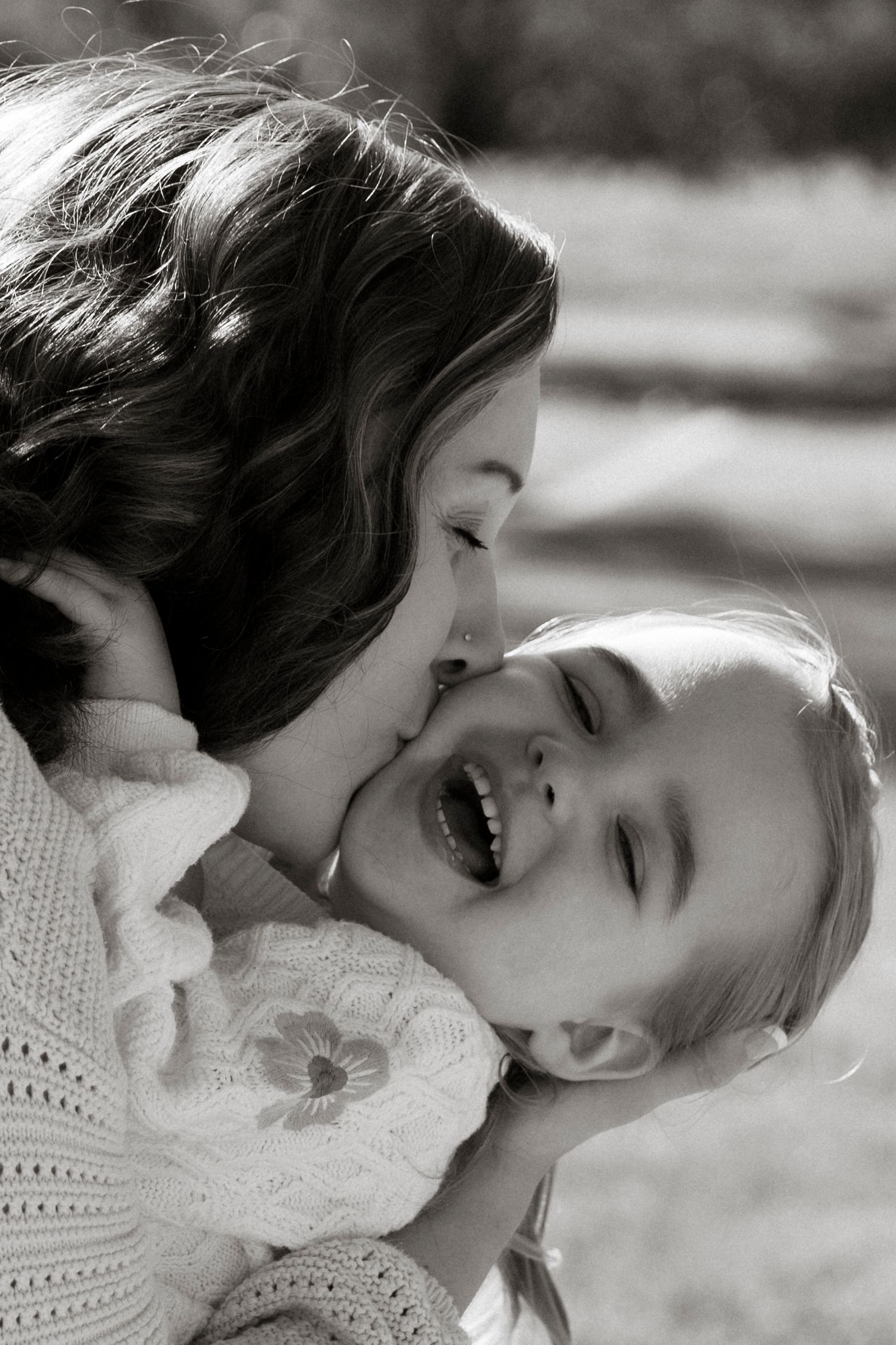 A black and white photo of a mother kissing her laughing young daughter on the cheek outdoors.Family photography in Regina Saskatchewan.