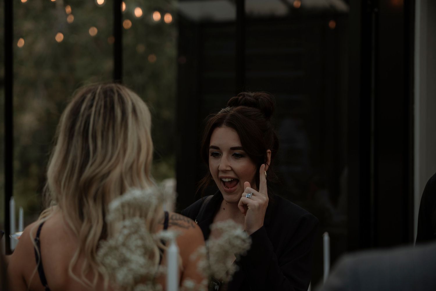 Two women laughing and talking at an outdoor evening dinner party with string lights in the background.