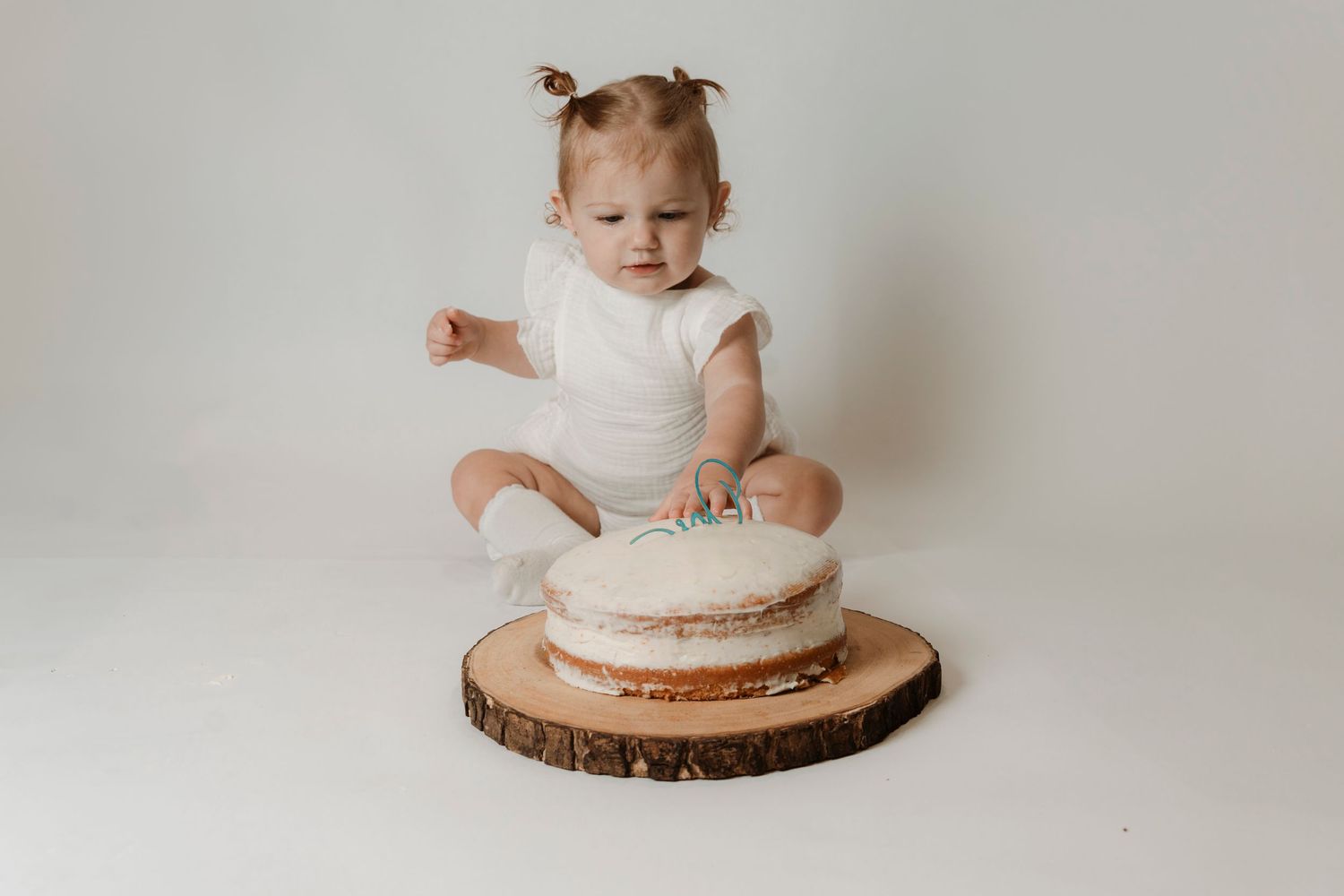 Baby girl in white romper sitting on floor smashing her first birthday cake on wood slice.Family photography in Regina Saskatchewan.