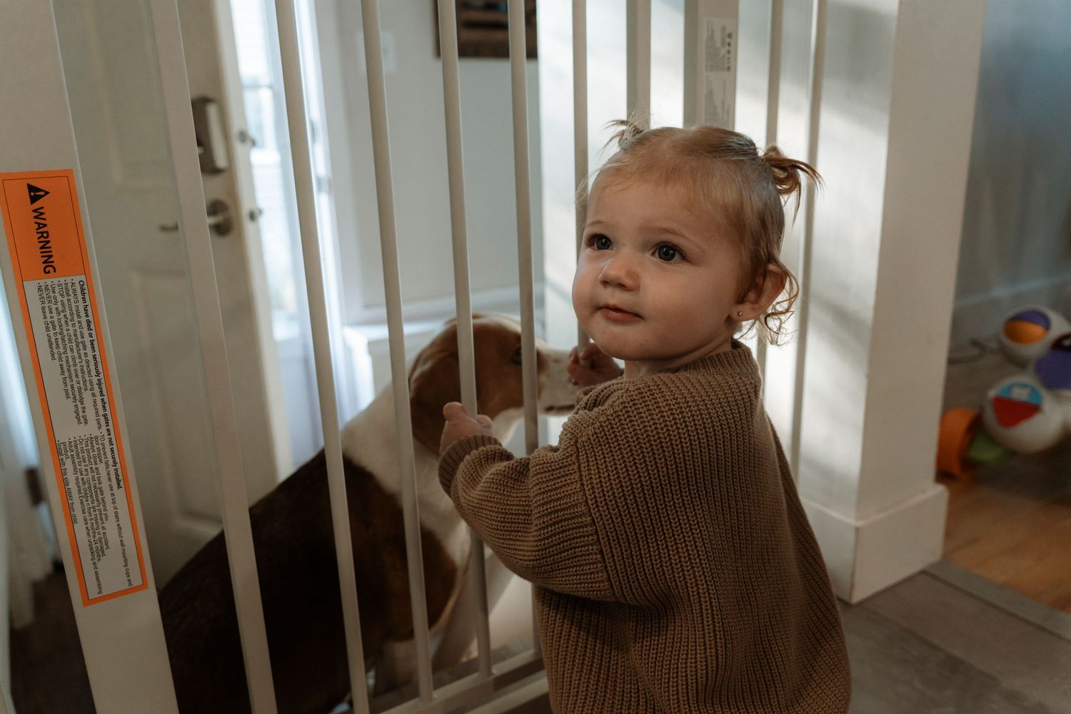 Smiling toddler girl in brown sweater standing at a white baby safety gate.Family photography in Regina Saskatchewan.