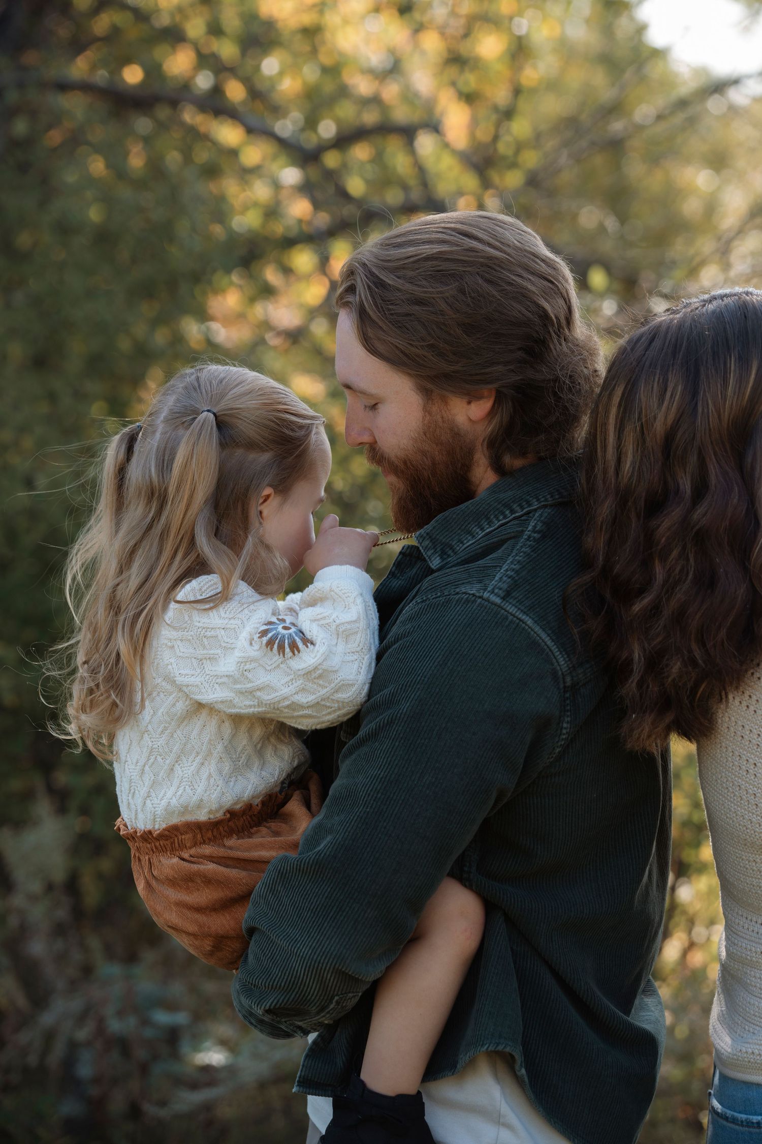 A bearded father tenderly holds his smiling toddler daughter close while standing outdoors among autumn foliage.Family photography in Regina