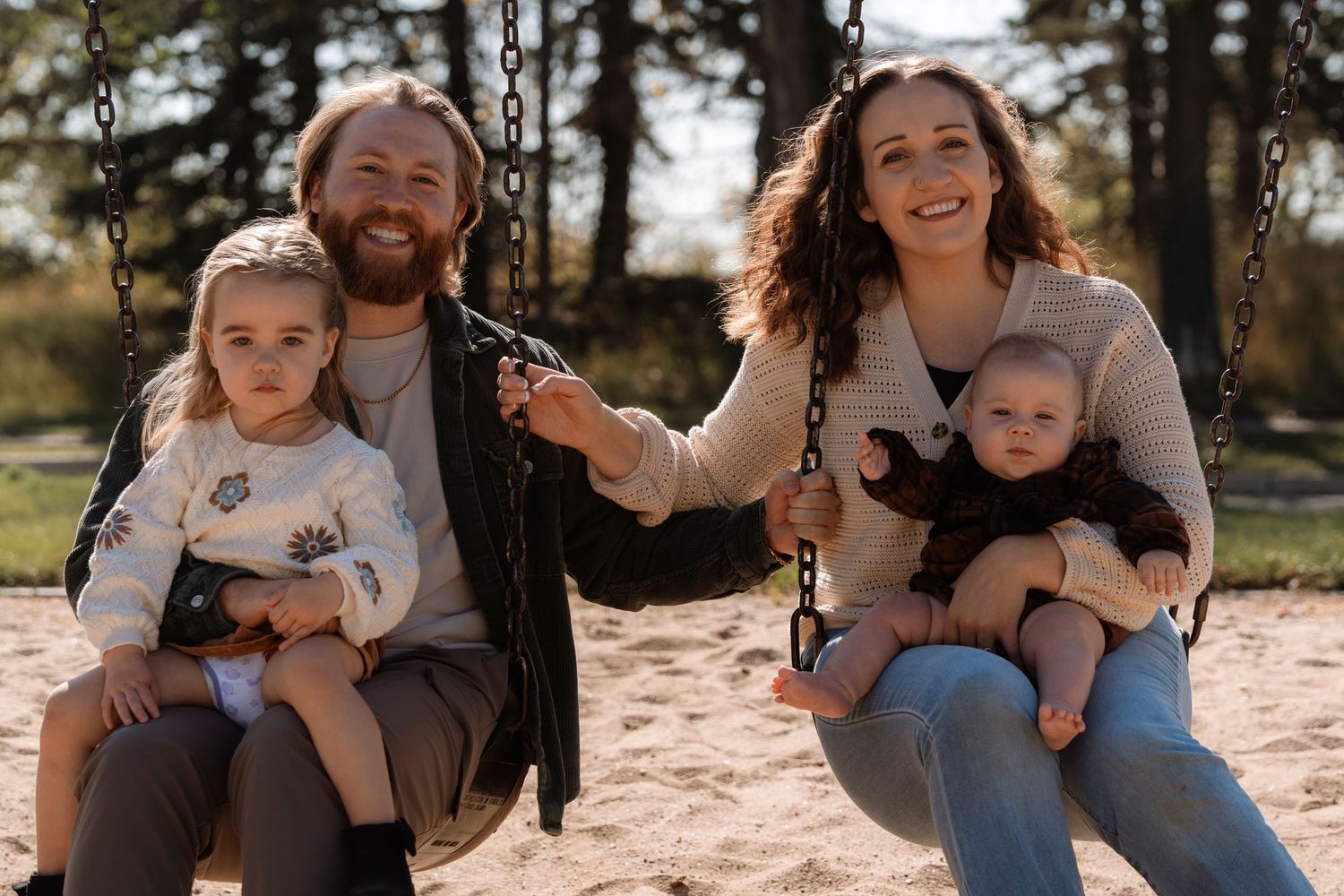 Happy family of four sitting on swings at a playground on a sunny autumn day, smiling at the camera. Family photography in Regina Sask.