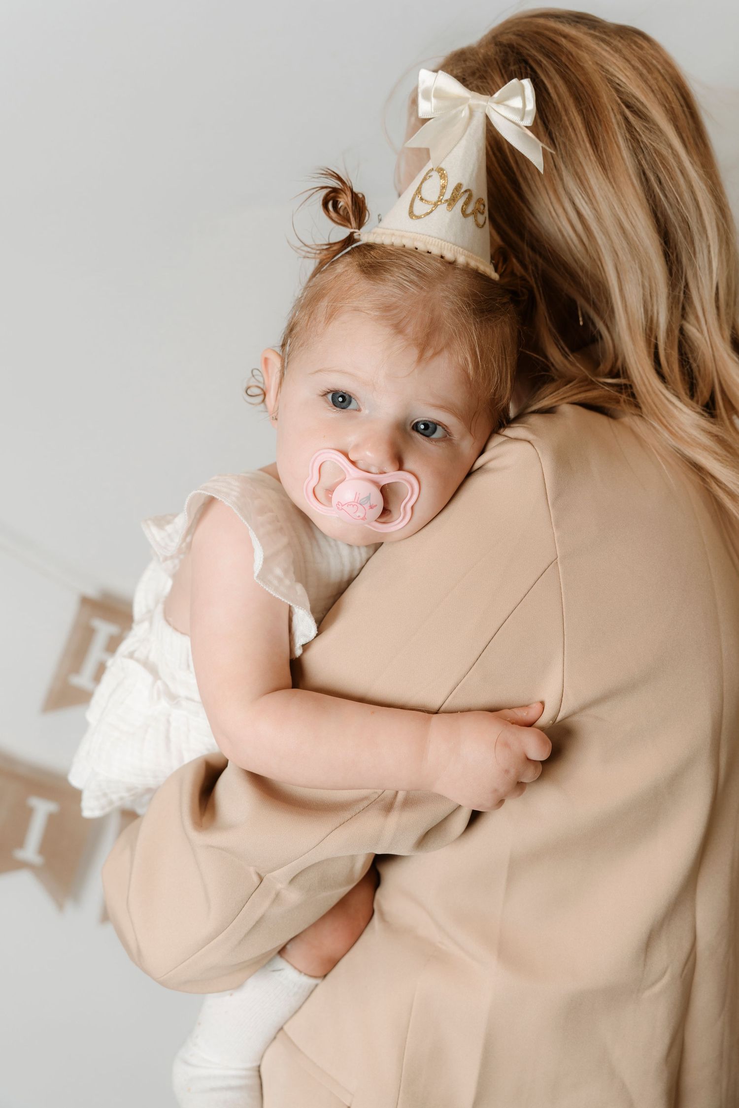 A smiling baby with a pink pacifier wearing a birthday crown is held by a woman in beige.Family photography in Regina Saskatchewan.