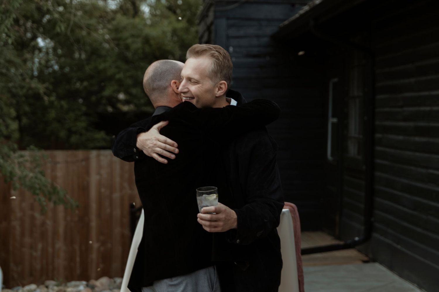 Two men in black sweaters share a warm hug outdoors near a wooden building, one holding a green drink.