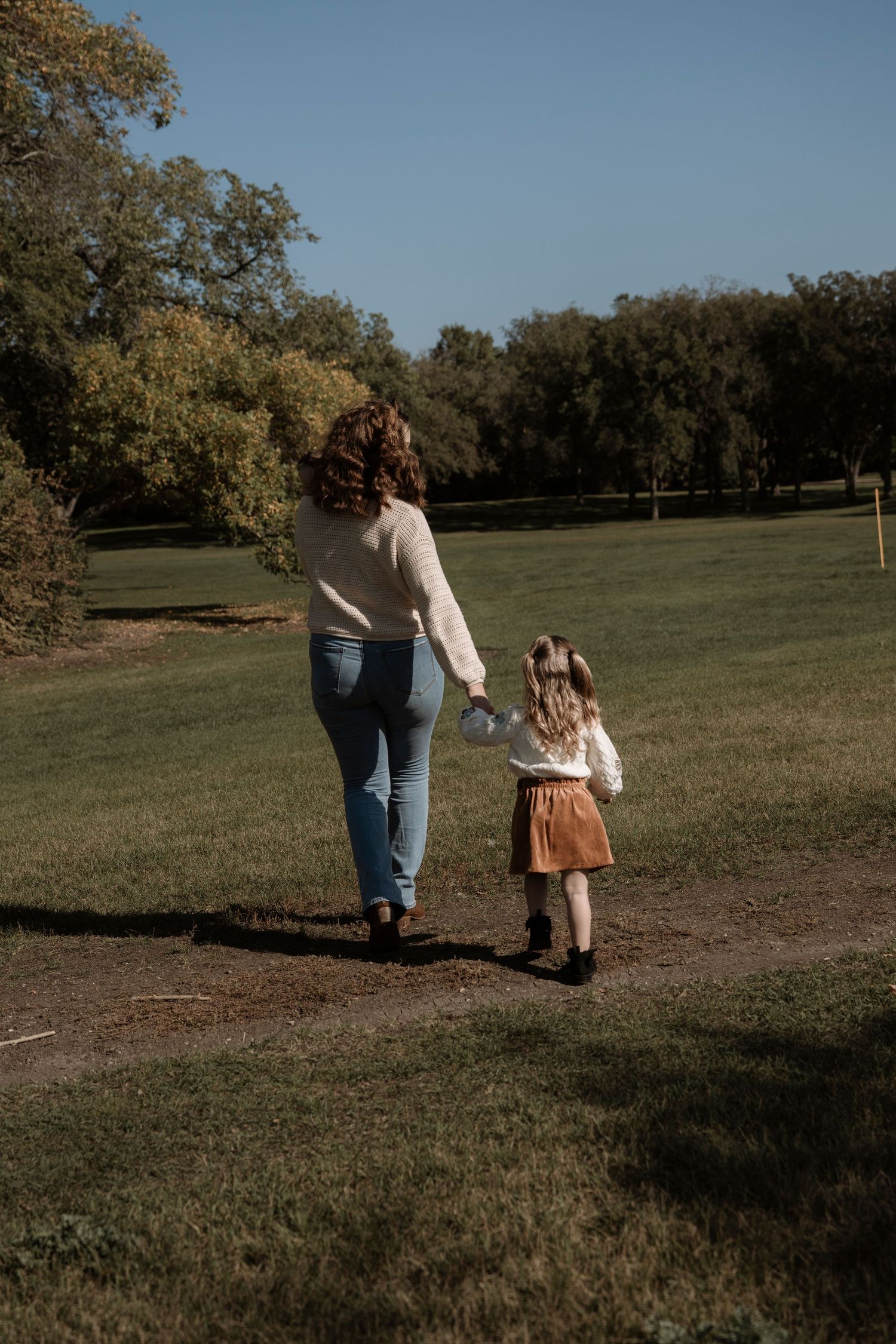 A mother and young daughter walk hand-in-hand along a path through a sunny green park on a clear day. Family photography in Regina Sask.