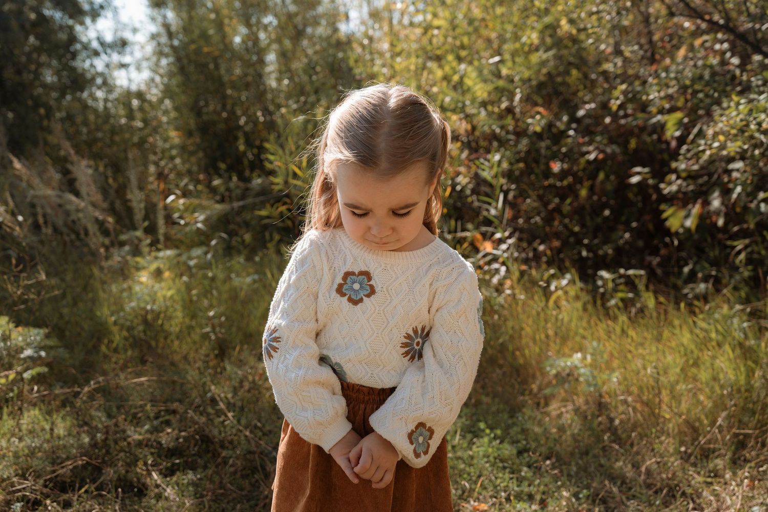 A young girl in a cream floral sweater and rust corduroy skirt looks down while standing outdoors.Family photography in Regina Saskatchewan.
