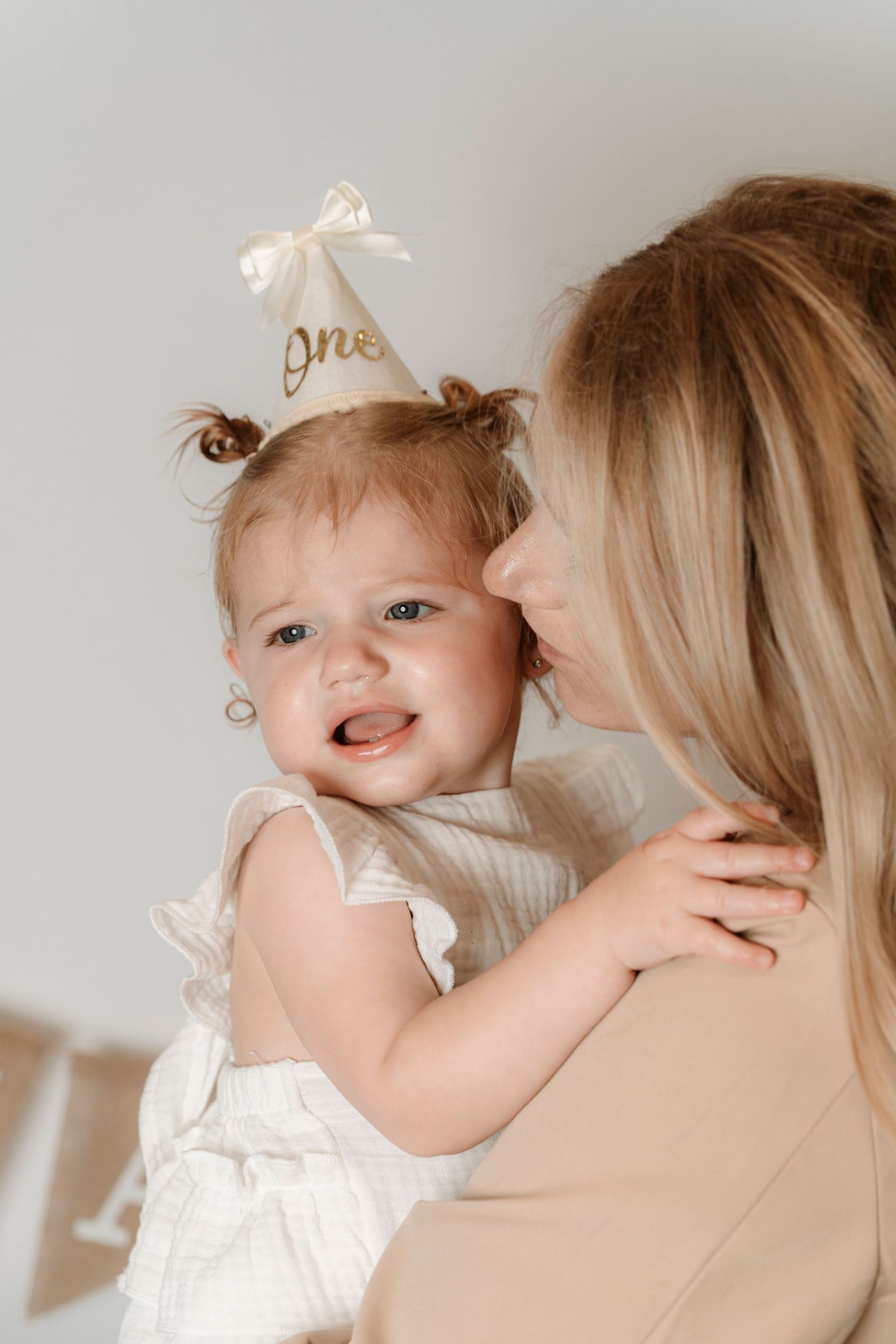 Mother kissing smiling baby girl wearing a white party hat on her first birthday. Family photography in Regina Saskatchewan.