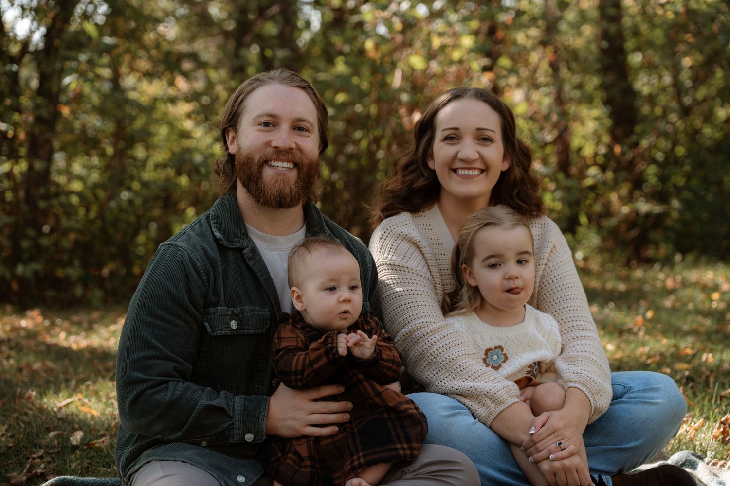 A smiling family of four sits outdoors in autumn, parents holding two young toddlers among colorful fall foliage.Family photography.