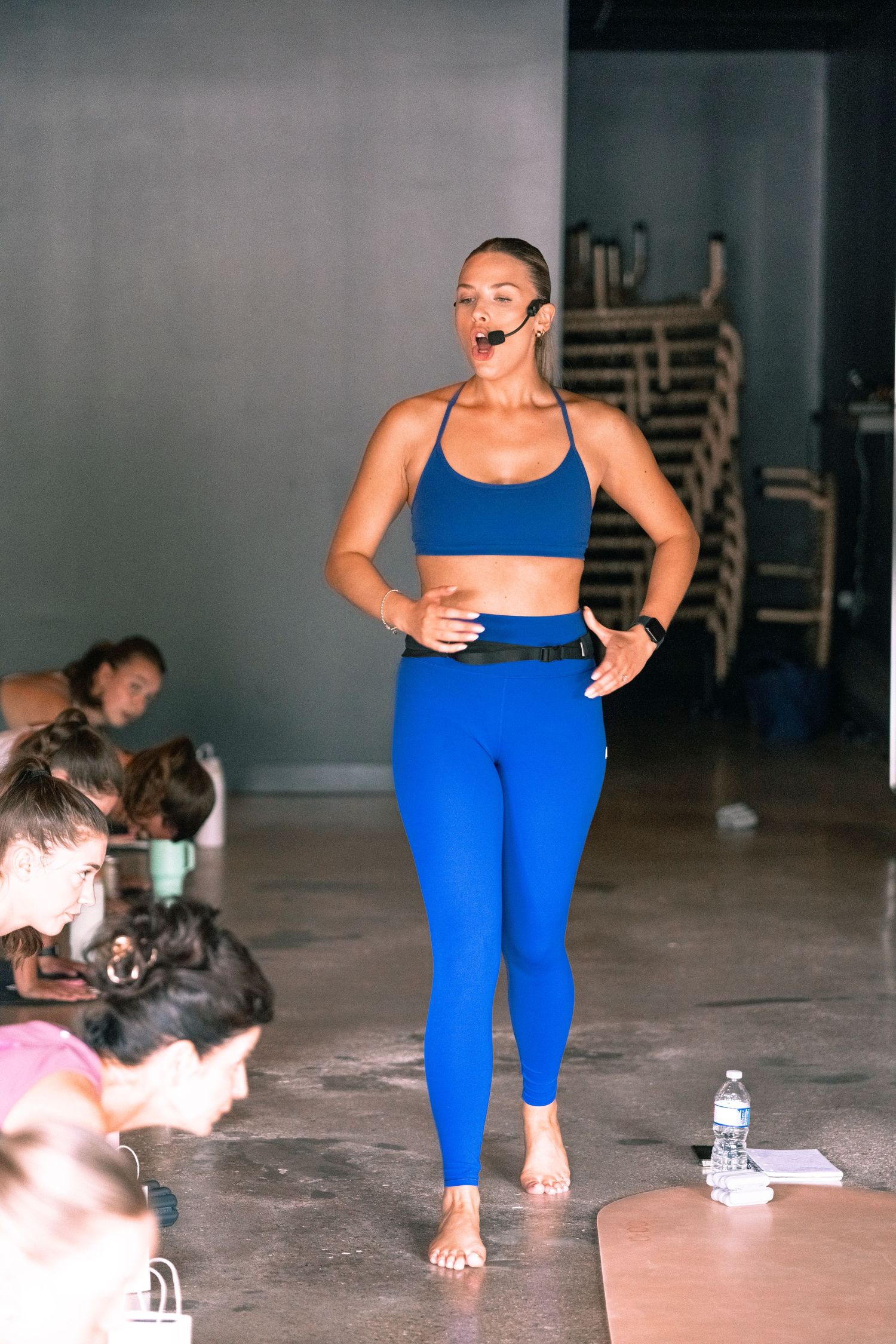 Woman in blue sports bra and leggings teaching a fitness class barefoot, wearing a headset microphone.