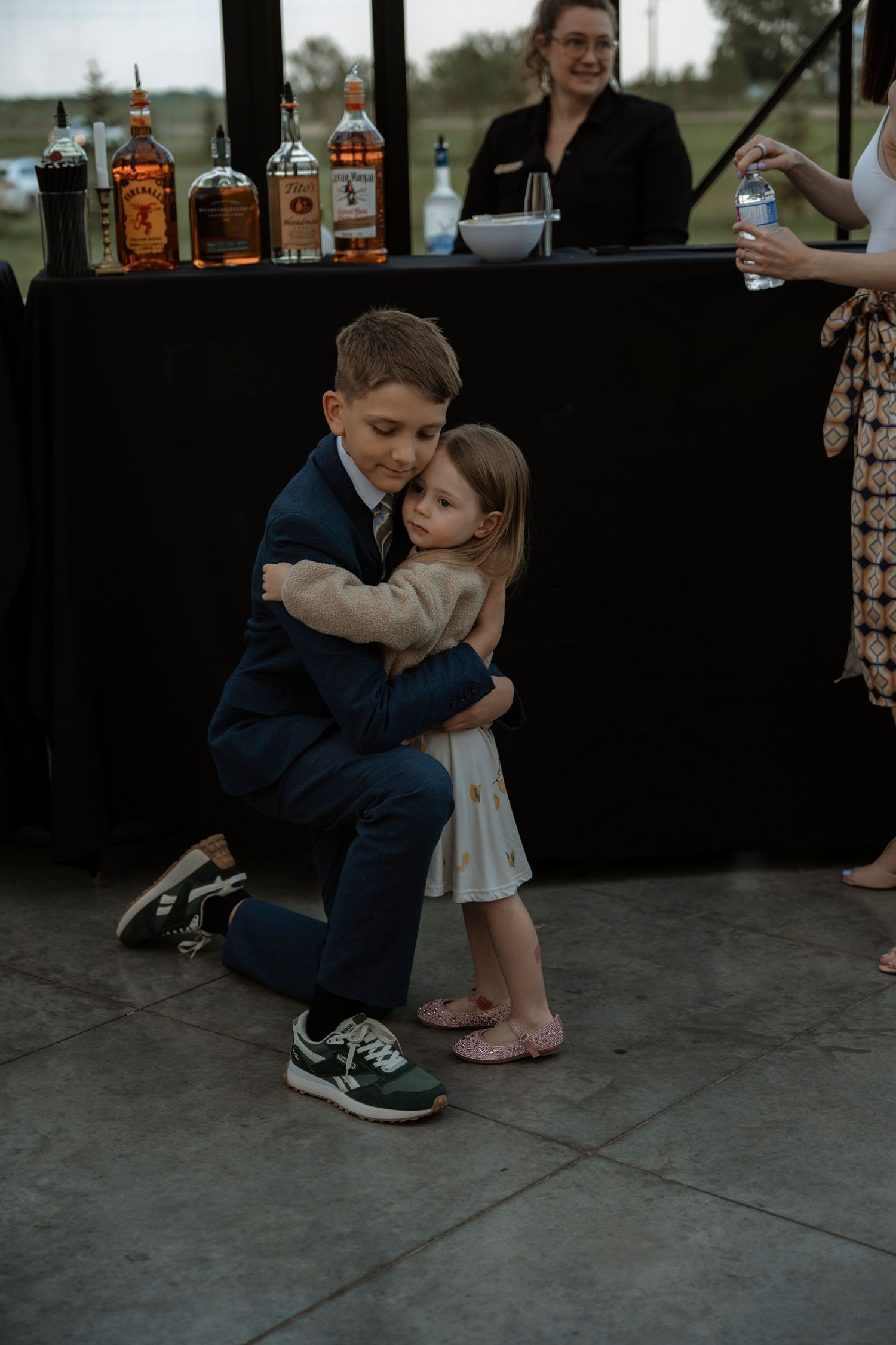 A boy in a navy suit kneels to hug a little girl in a white dress at a wedding reception.
