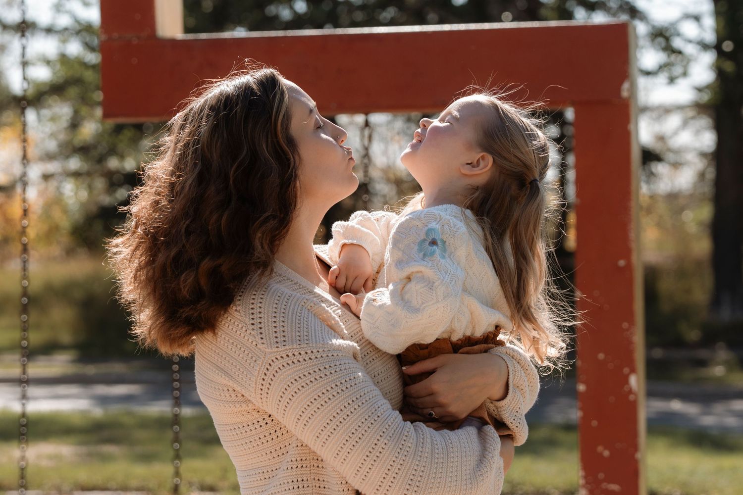 A curly-haired mother lovingly holds her young daughter near a playground swing set in warm sunlight.Family photography in Regina Sask.