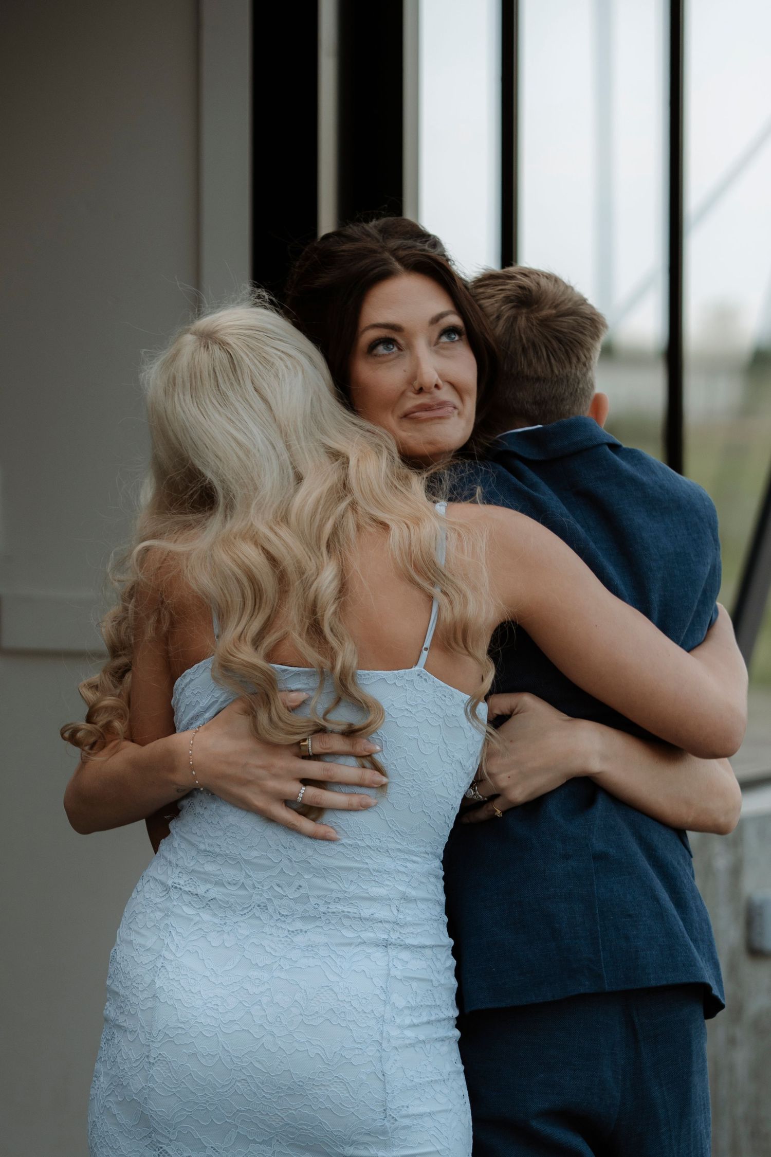 Emotional woman with dark hair hugging two people, wearing a light blue dress at a formal event.
