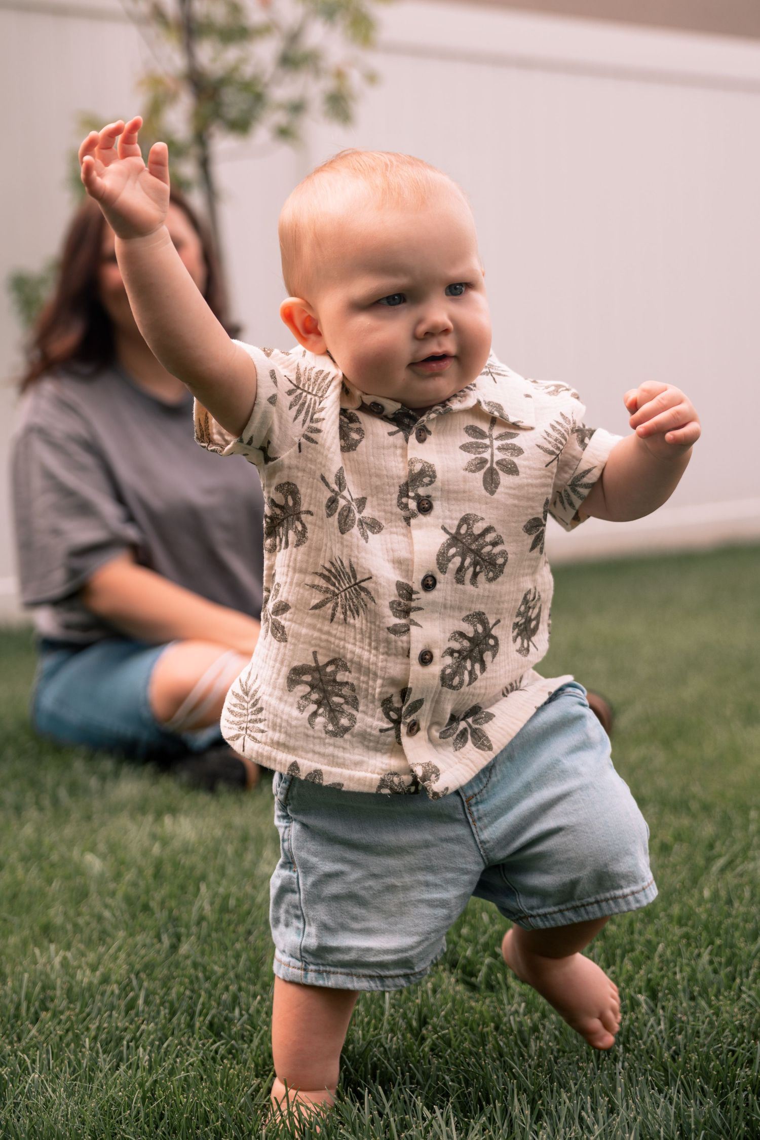 A baby boy in a floral shirt and denim shorts takes his first steps on green grass with a woman sitting behind him.