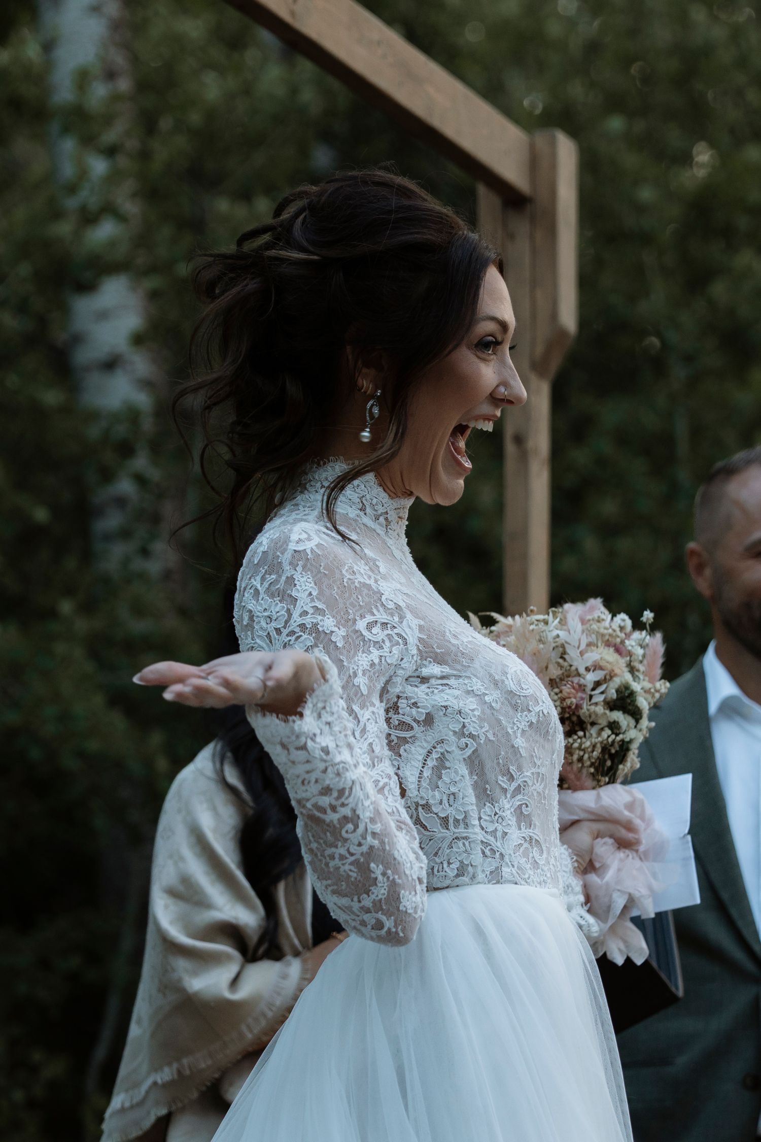 Bride in lace long-sleeve wedding dress laughing joyfully while holding a bouquet during an outdoor ceremony.
