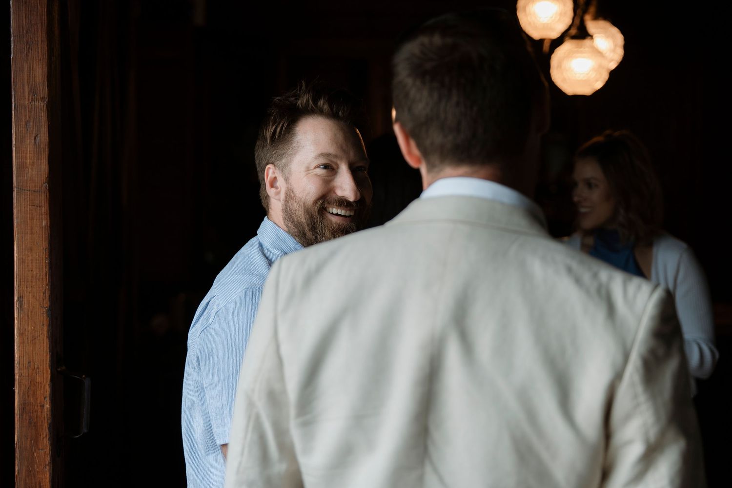 Two men smiling and conversing at an indoor event, with warm pendant lights and a woman visible in the background.
